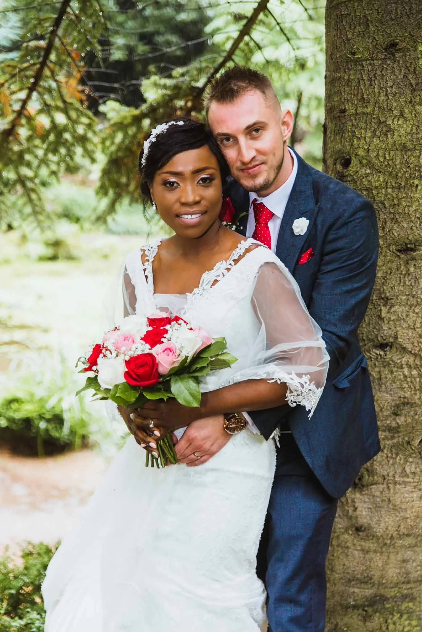 An interracial couple with males back to a tree holding the bride with a bouquet lookig at the camera, smiling.