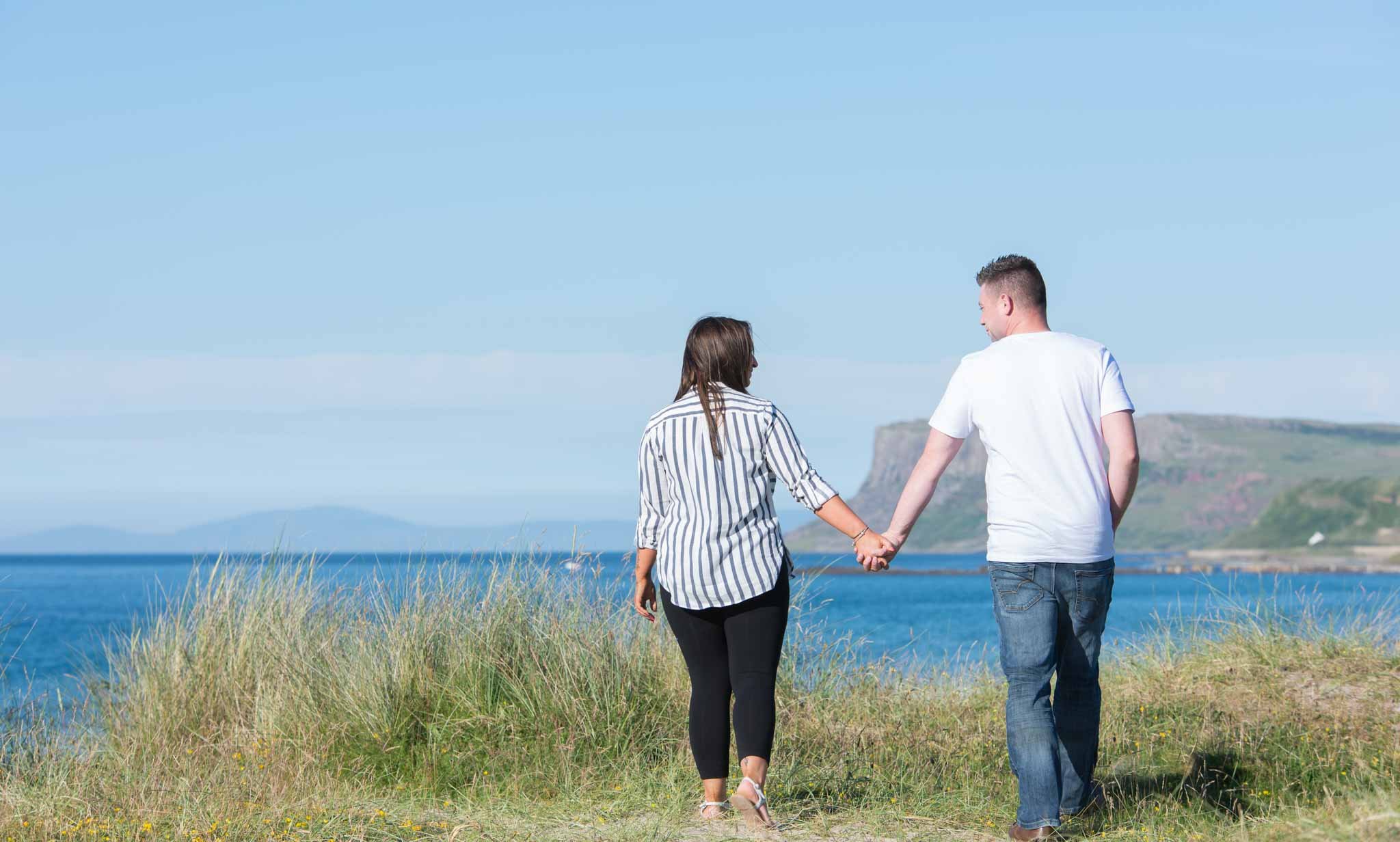 An engaged couple walking along a north coast beach of Northern Ireland, holding hands with the sea and cliffs in the distance.