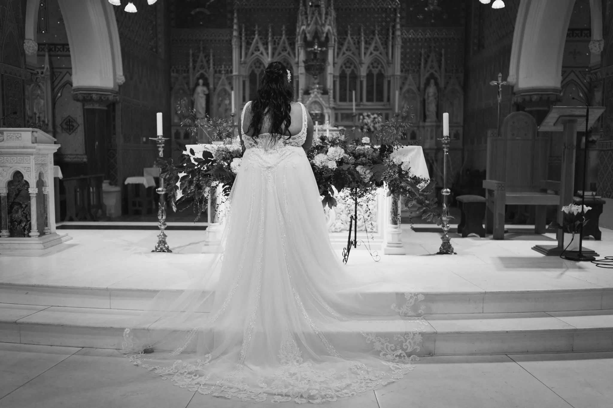 Mono image of the back of brides dress at the front of the church with lighting from large windows.