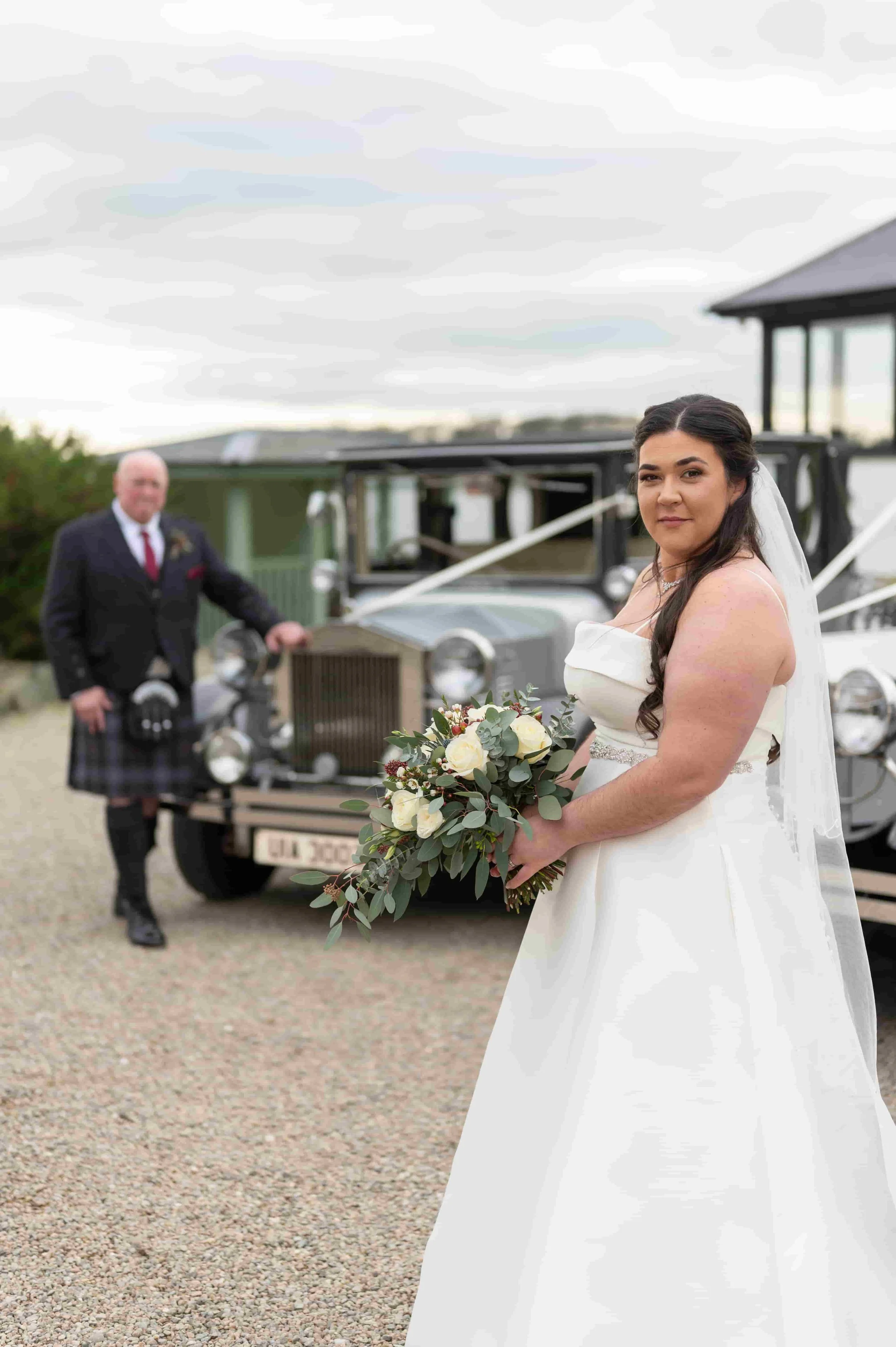 Bride holding a bouquet with the groom in the distance.