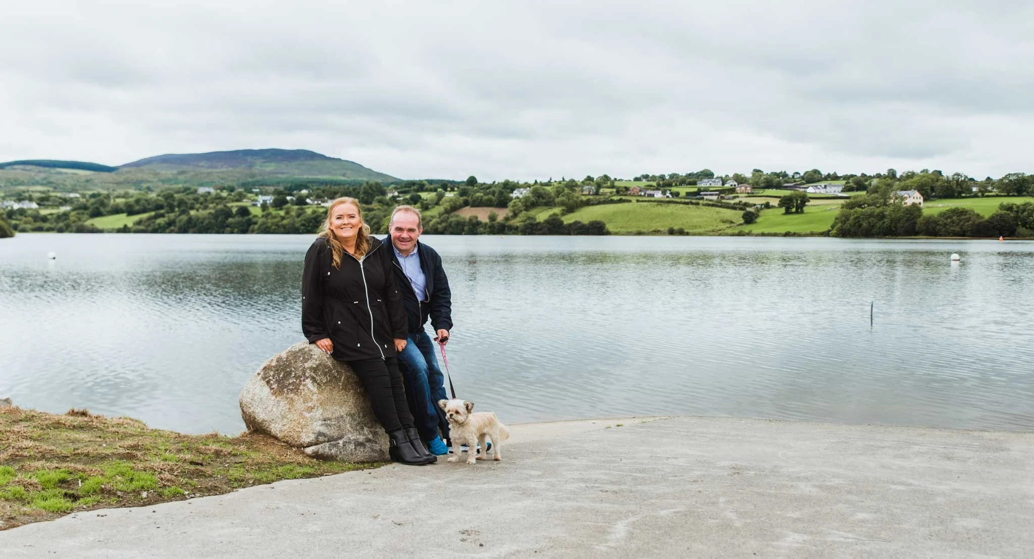 The couple is sitting at a rock with the lake and countryside as a backdrop.
