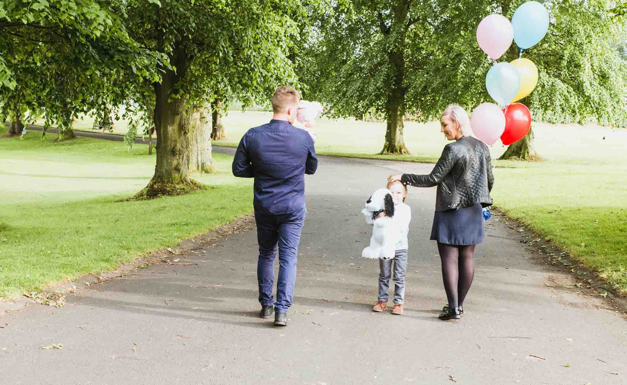 Two adults and a kid with a dad holding a baby walking along a path. Mum is holding balloons.