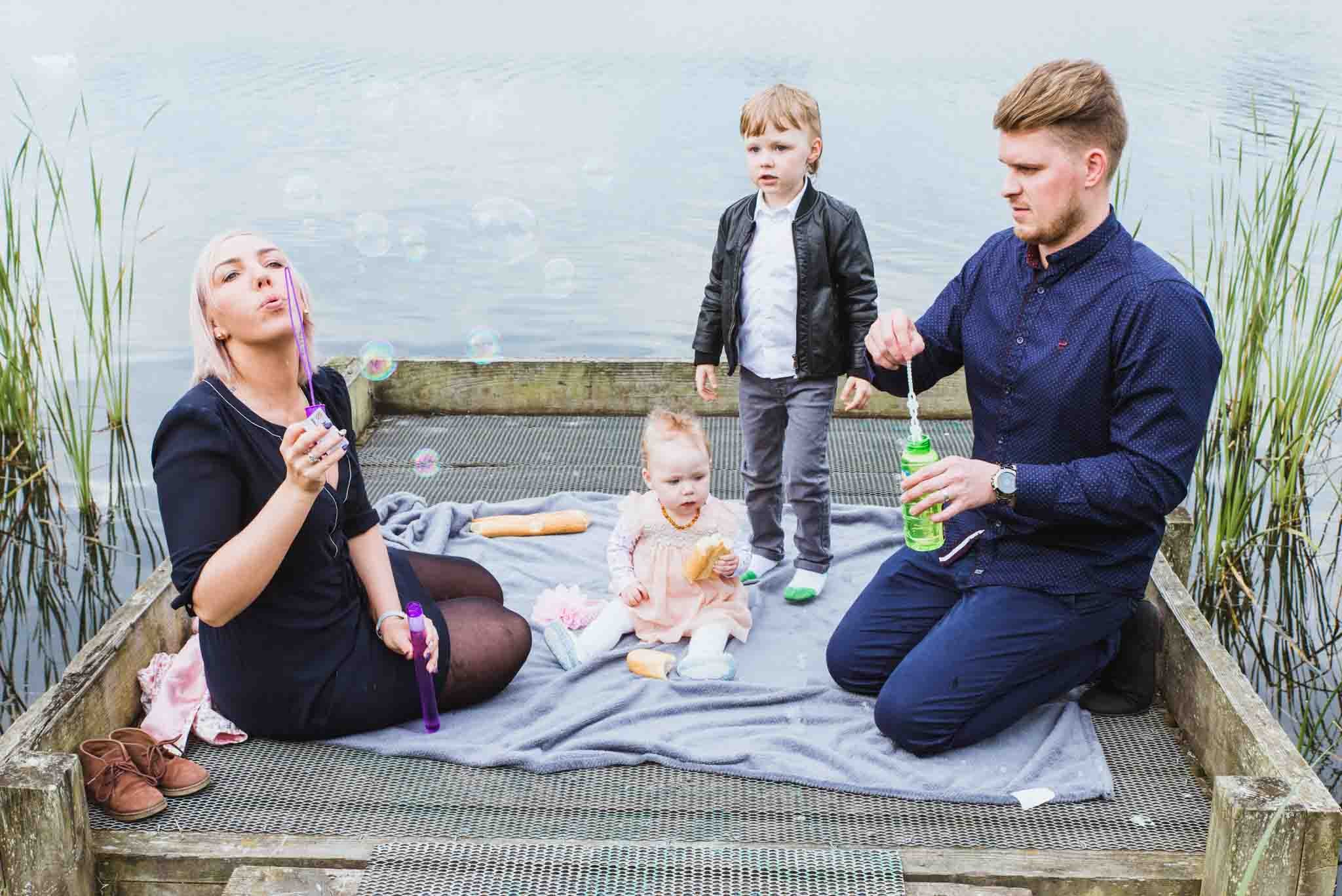 The family is sitting down on the jetty as the mother blows bubbles.