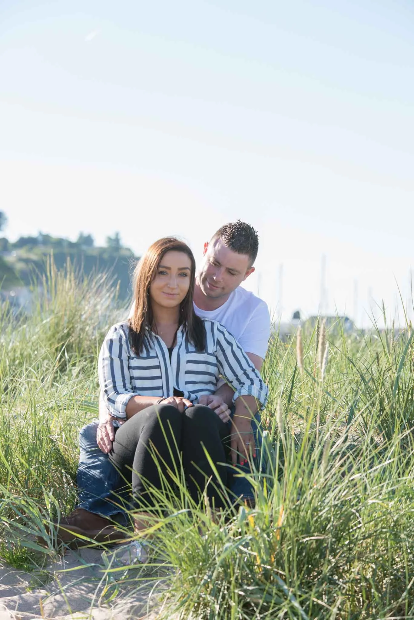 A male sitting behind a female as she smiles, looking at the camera on a beach with cliffs and a clear blue sky in the background.