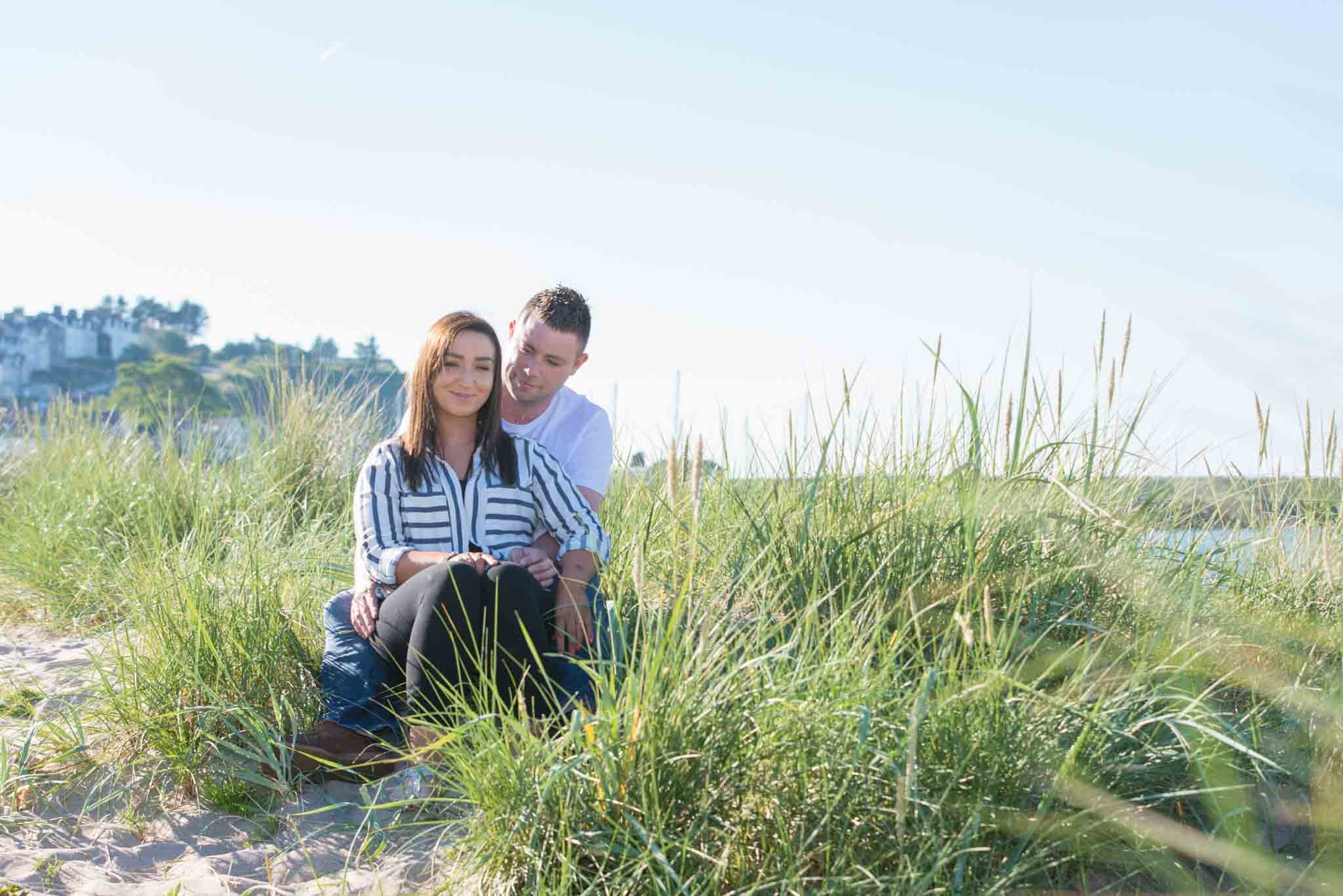 A couple is sitting down on the long grass at the beach.