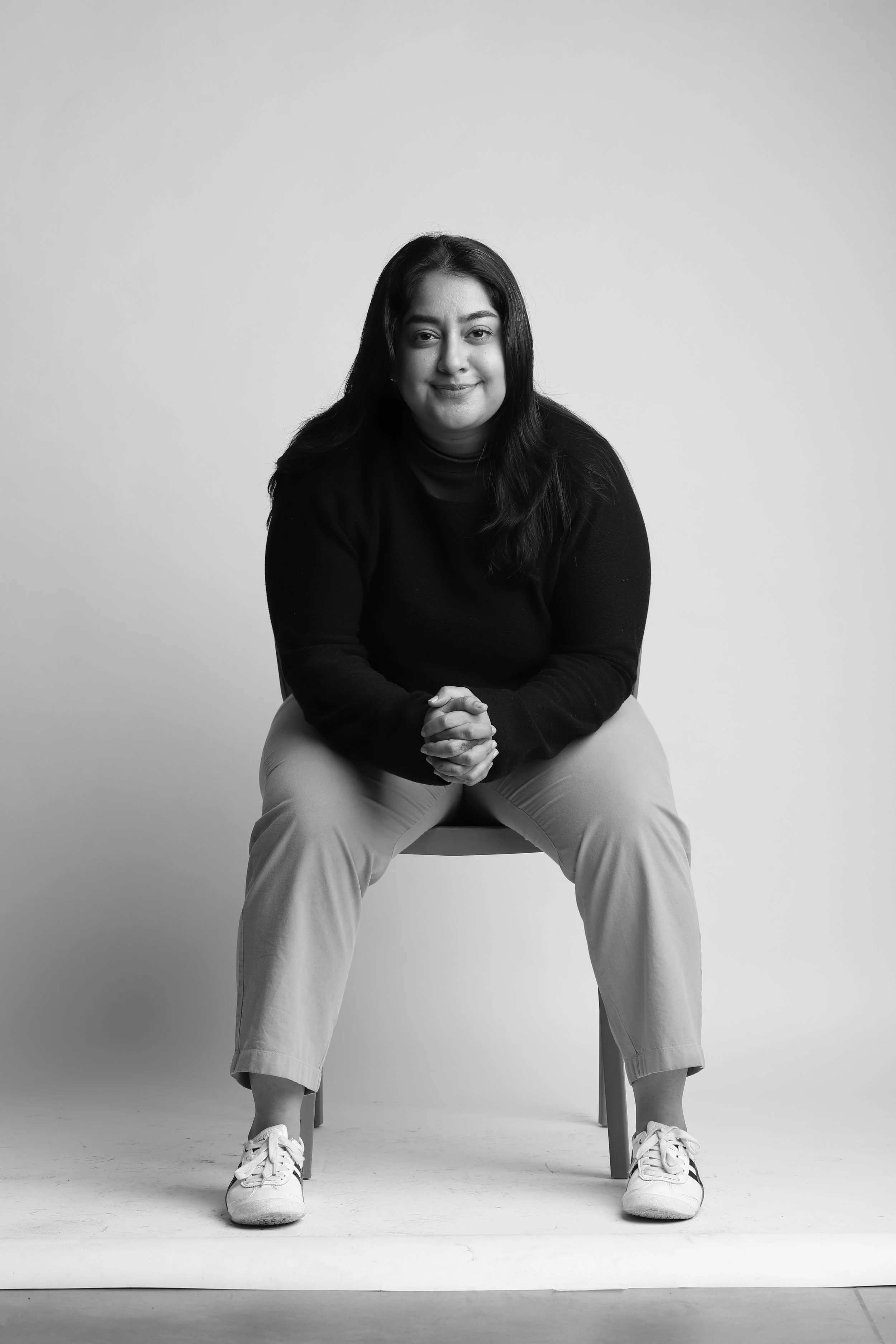 Black and white portrait of a woman sitting on a chair, smiling, with hands clasped, wearing a dark top, light-colored pants, and sneakers.