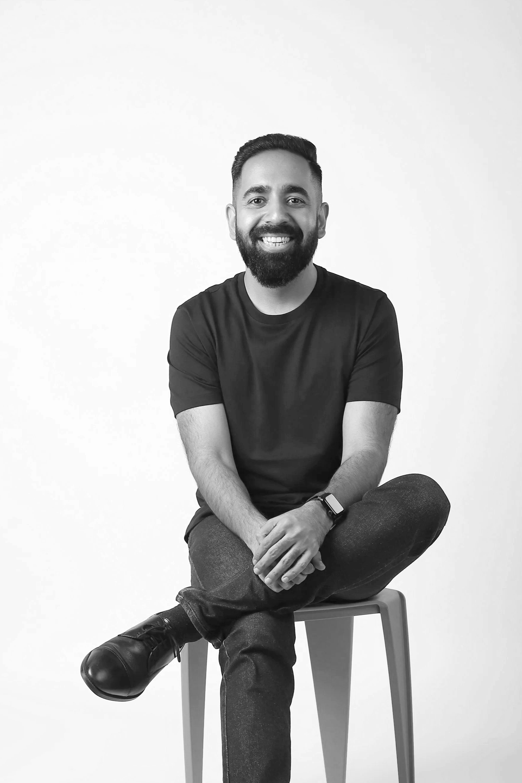 A smiling man with a beard and tattoos, sitting cross-legged on a chair against a plain background, wearing a black t-shirt and jeans.