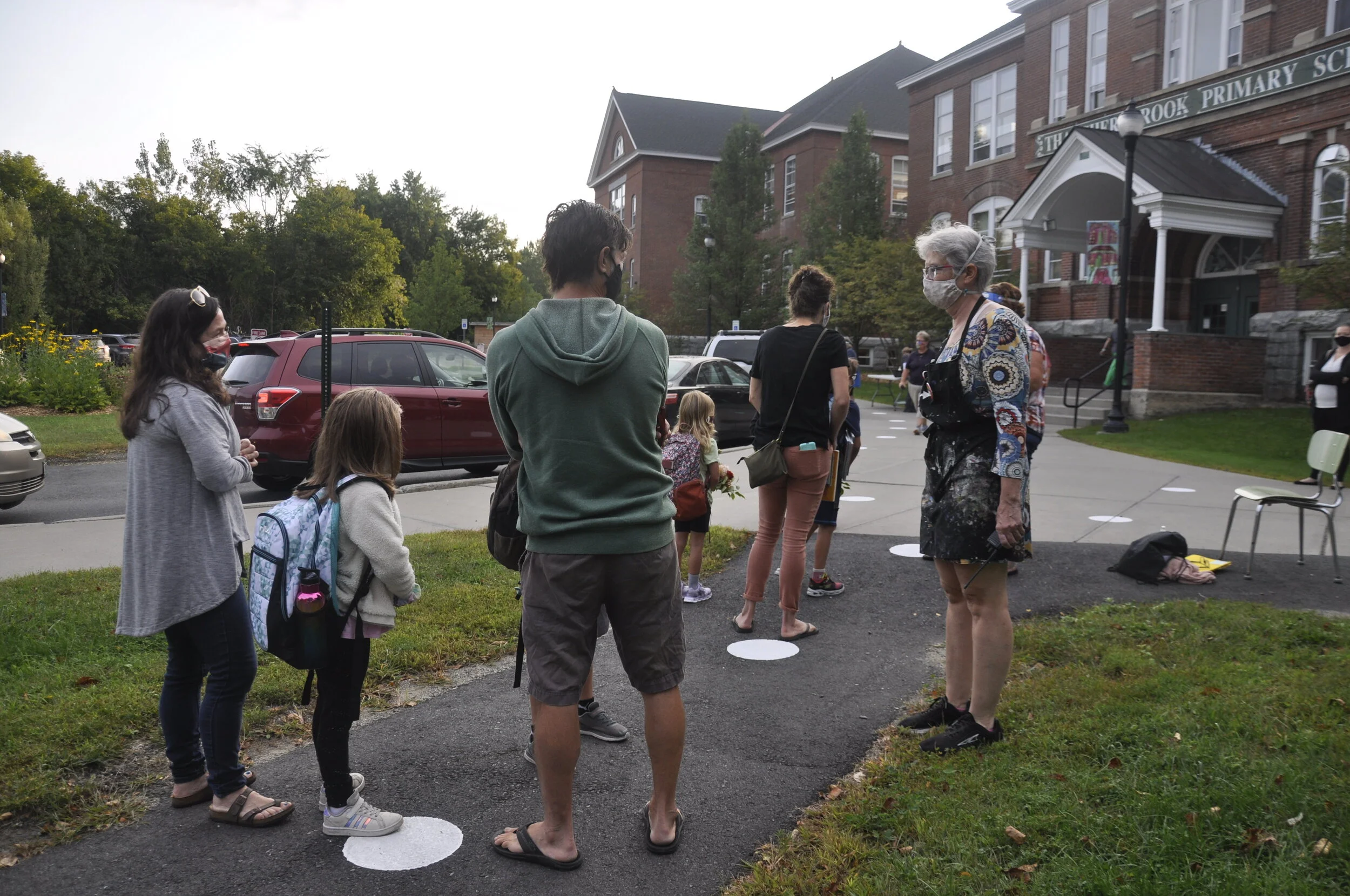    Art teacher MK Monley (right) visits with students and families in line. The new white dots painted on the sidewalks around school help everyone with the distancing routine.  
