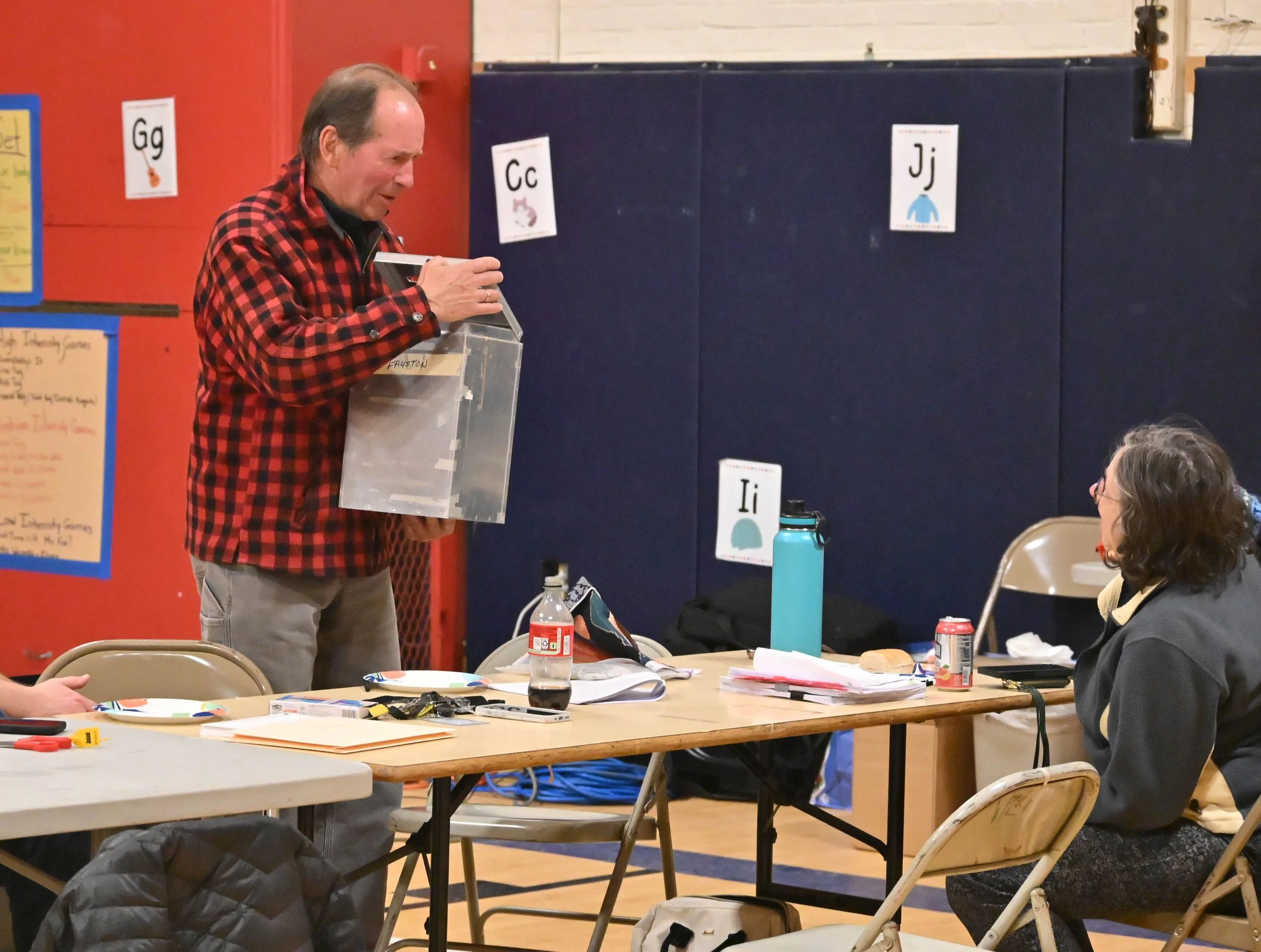   Jared Cadwell arrives from Fayston with a box of Harwood school district election ballots to be counted at Brookside Primary School on Tuesday night, with Waitsfield election official Bobbi Rood (seated). Photo by Gordon Miller  