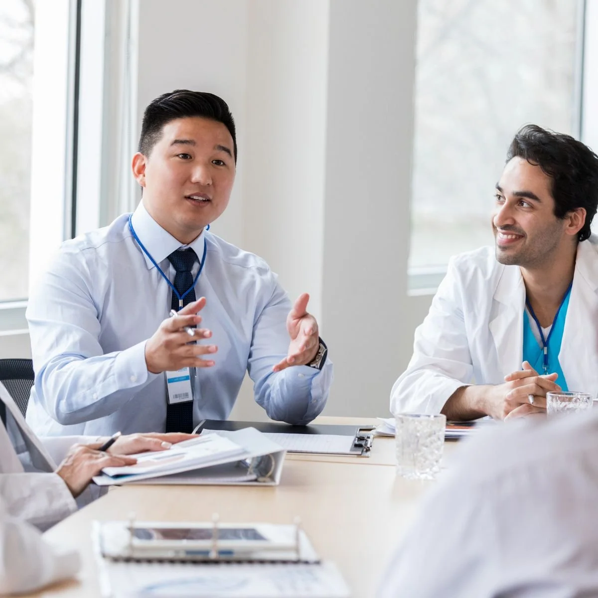 Two male doctors engaged in a discussion at a conference table, with one speaking and the other listening, in a well-lit room with windows.