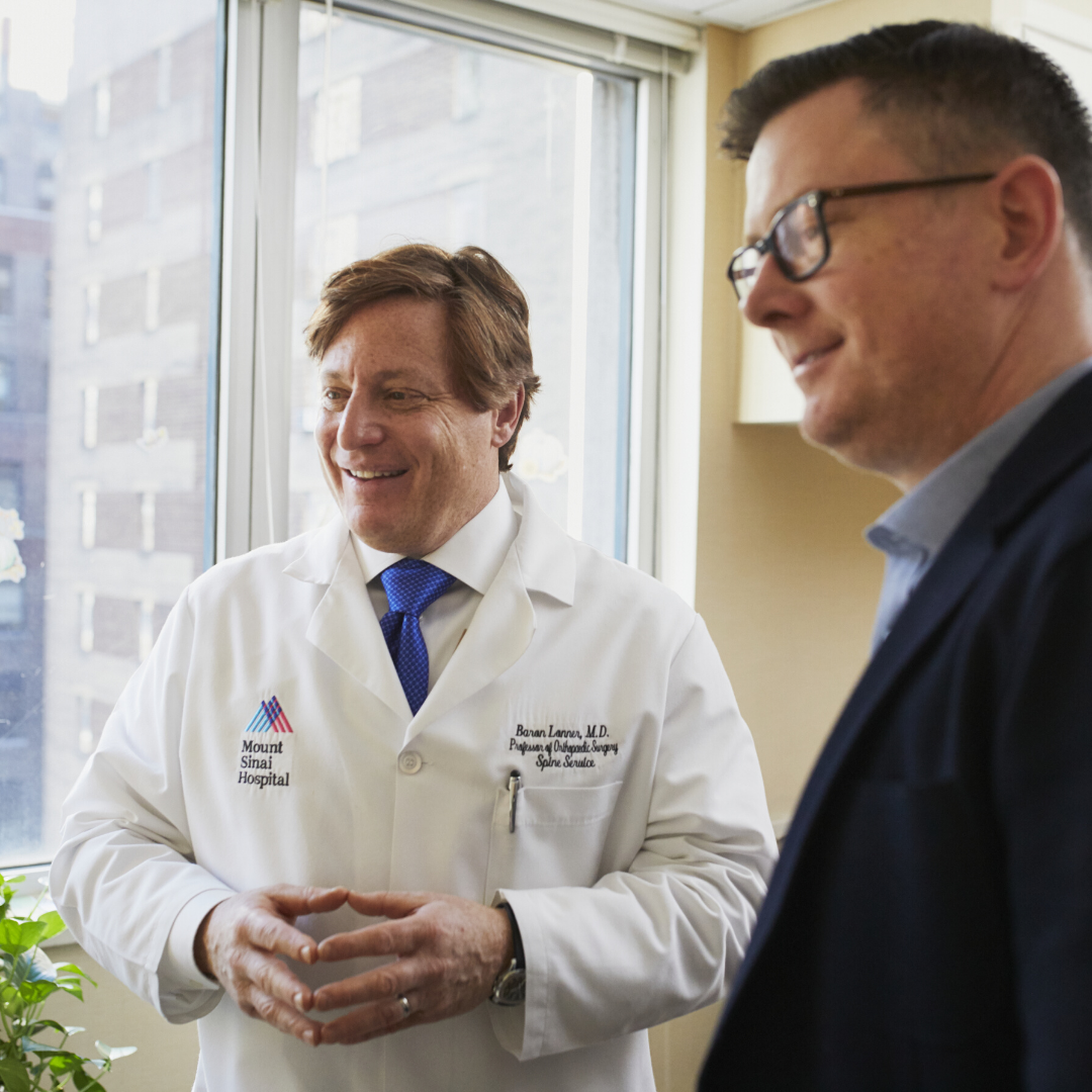 Two men standing indoors, one in a white lab coat with "Mount Sinai Hospital" logo, smiling near a window.