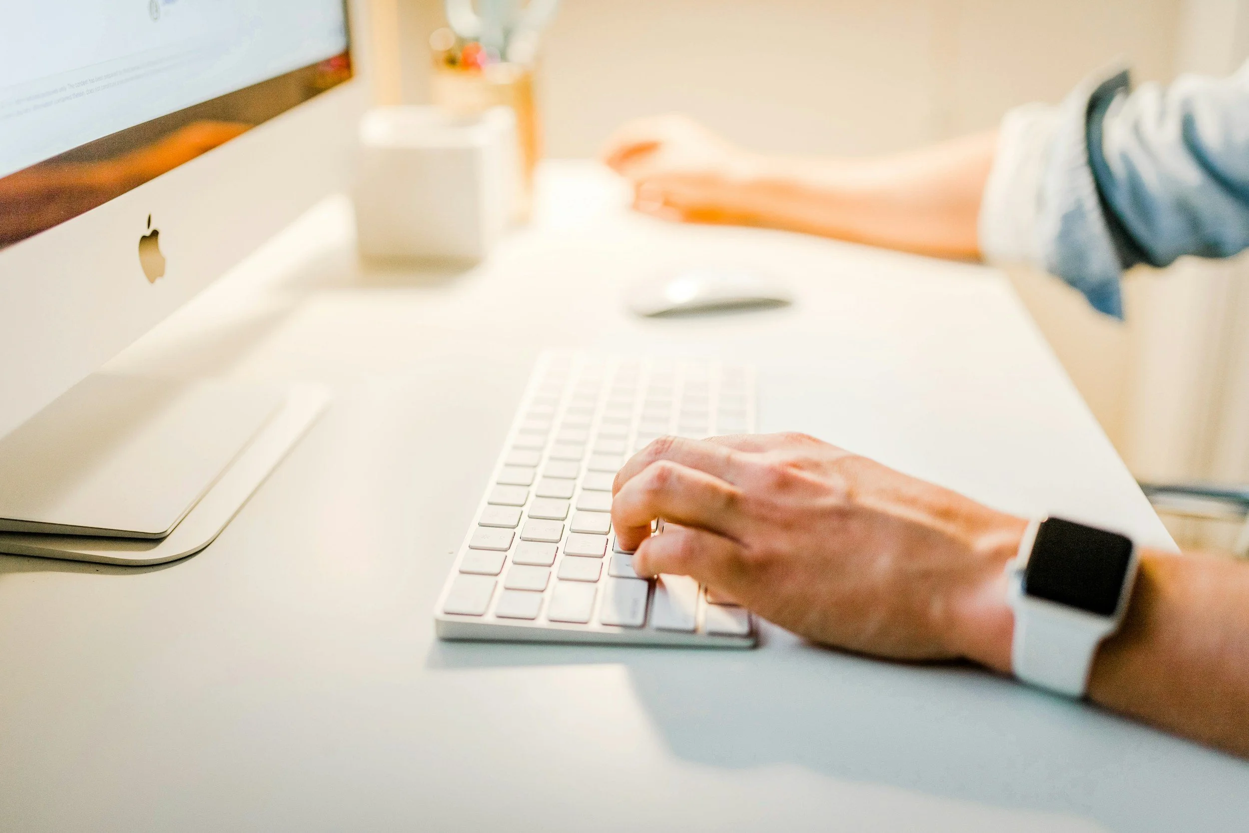 Person typing on an Apple keyboard, using a mouse, with a smart watch on the wrist, near an Apple iMac.