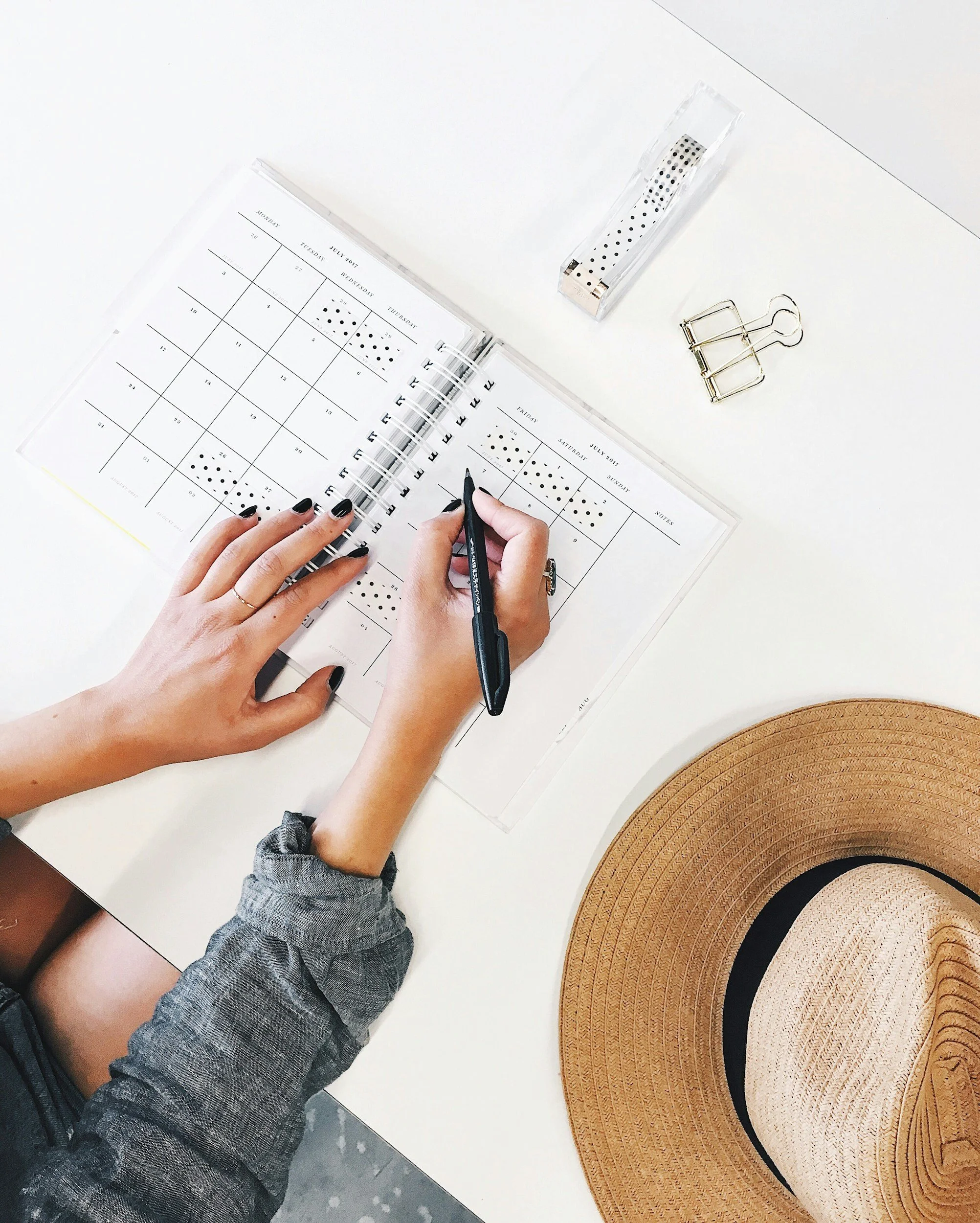 Person writing in planner, wearing a gray shirt, with a hat, binder clip, and tape dispenser on the desk.