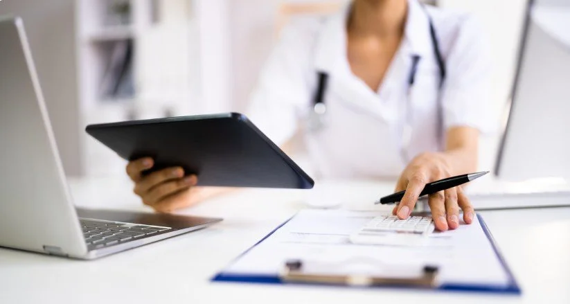 Medical professional in white coat using a tablet and calculator at a desk with a laptop.