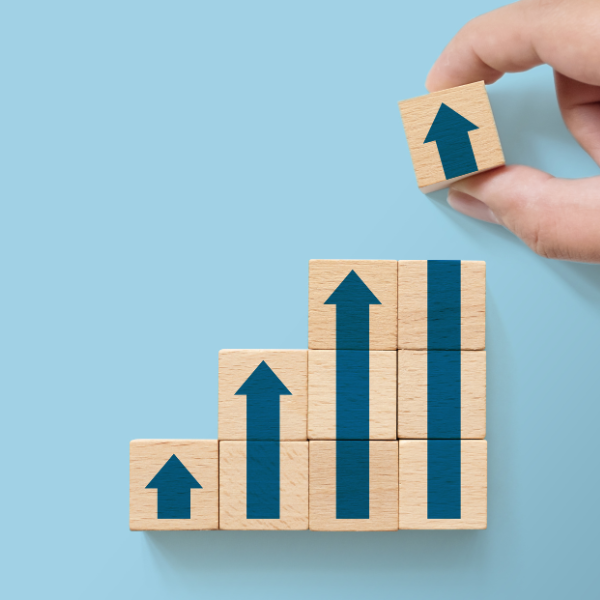 Hand placing a wooden block with an upward arrow on top of a staircase of blocks with upward arrows, symbolizing growth or progress.