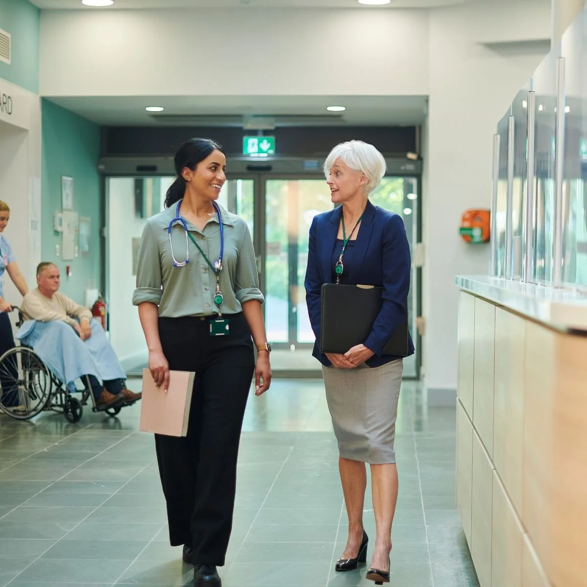 Two female medical professionals walking and talking in hospital corridor, with elderly patient in wheelchair in background.