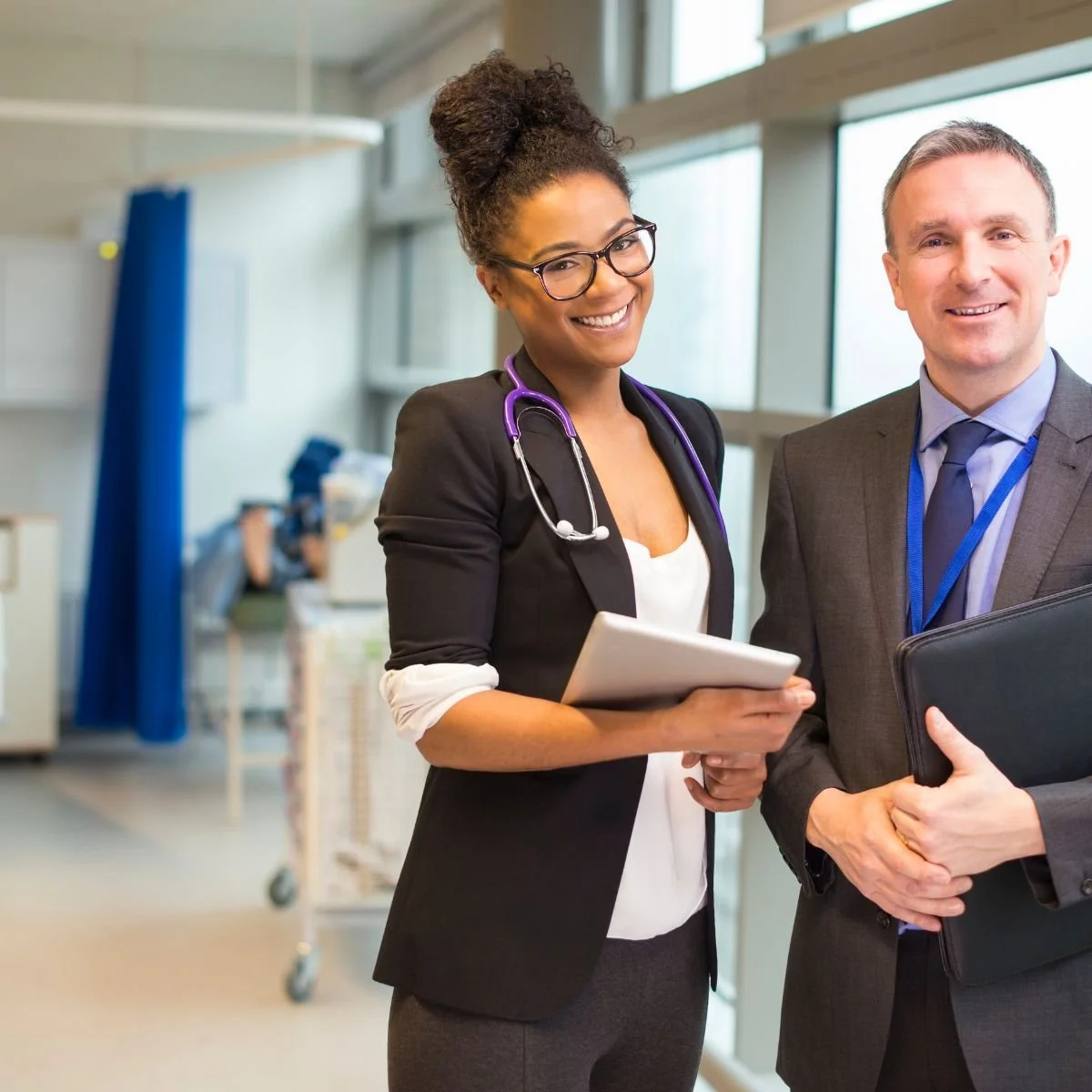 Two medical professionals, a woman with glasses and a stethoscope around her neck, and a man in a suit, smile and pose for the camera in a hospital or clinic setting.