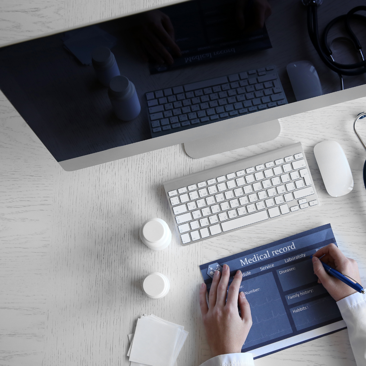 A healthcare professional filling out a medical record form on a white desk with a desktop computer, keyboard, mouse, bottles, and medical supplies visible.