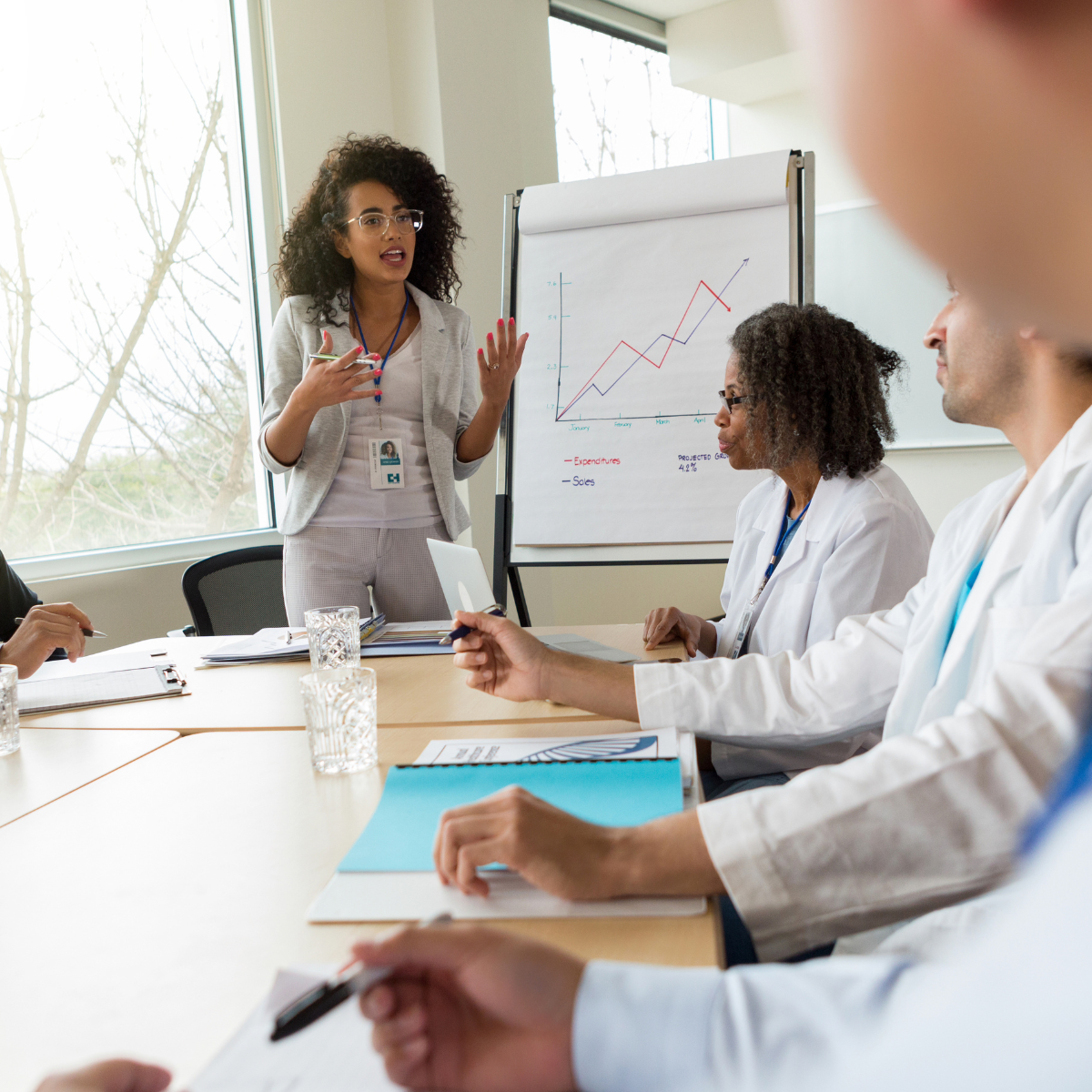 A woman giving a presentation in a meeting room with a flip chart showing a line graph of expenditures and sales, while three colleagues, two women and one man in lab coats, listen attentively.