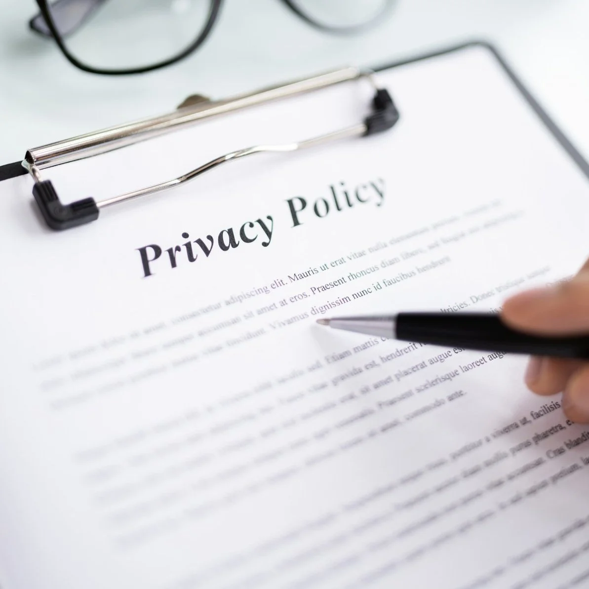 Close-up of a hand holding a pen near a printed 'Privacy Policy' document on a clipboard, with glasses in the background.