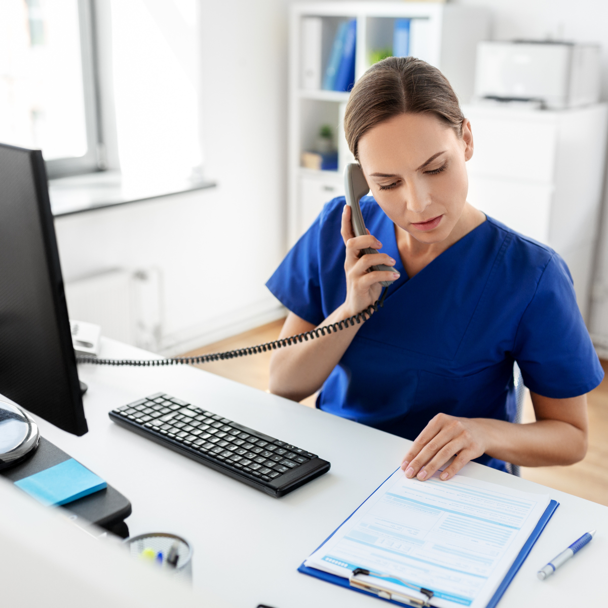 A nurse in blue scrubs talking on the phone at a desk in a medical office, with a computer, keyboard, and medical documents on the desk.