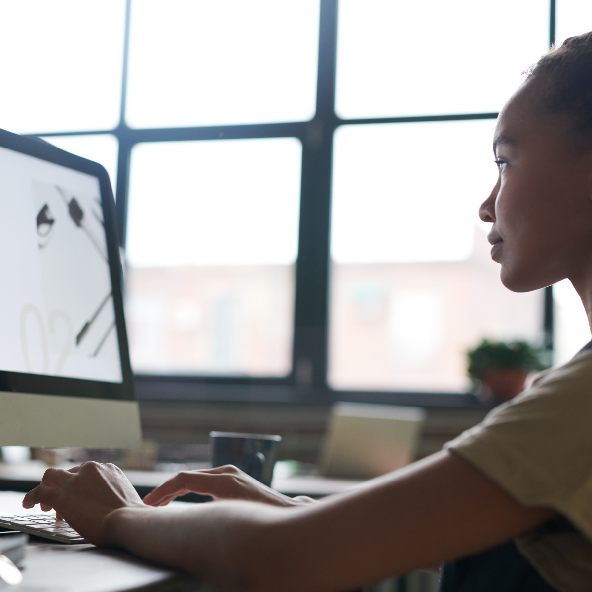 A woman working at a computer in a bright office with large windows.