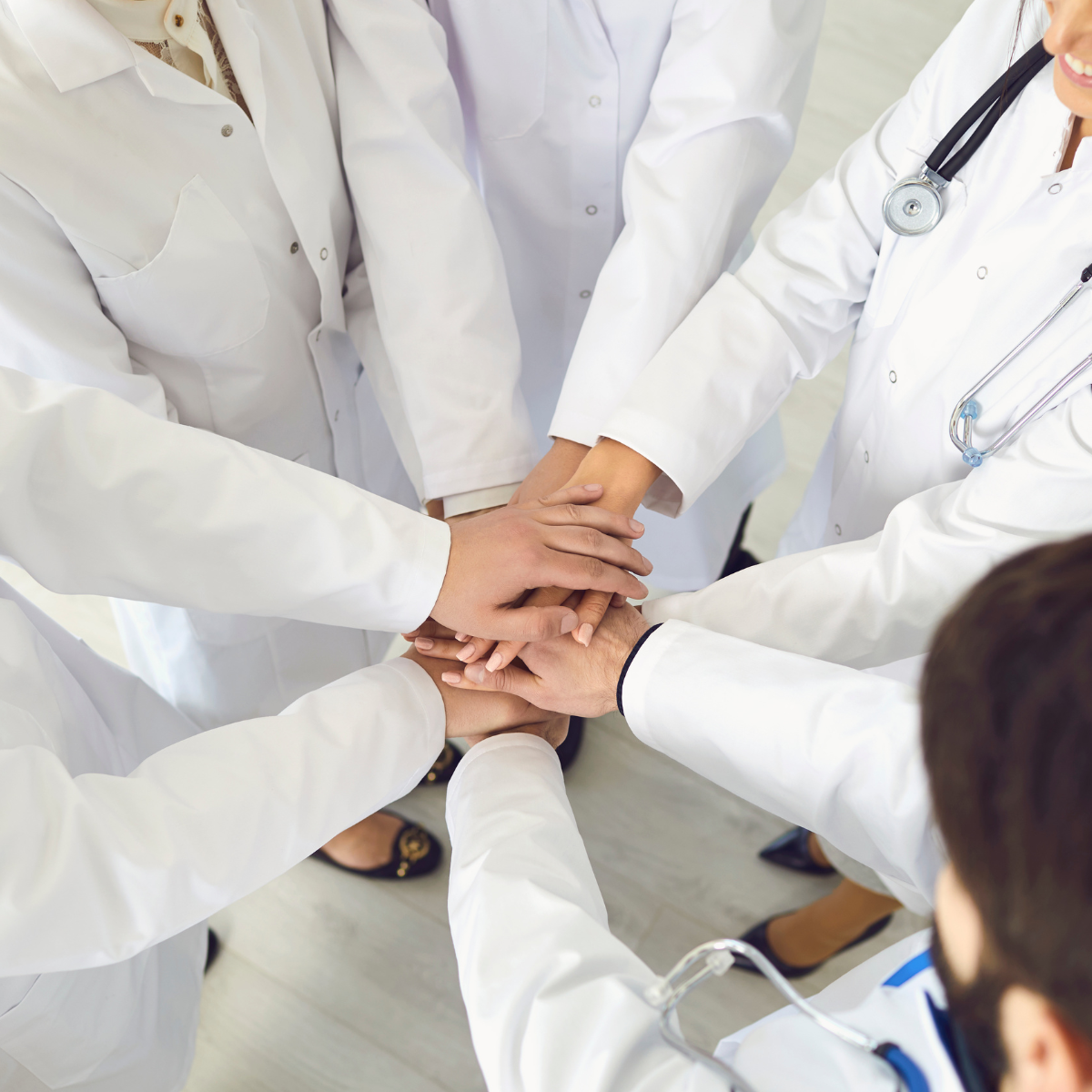 Group of medical professionals in white lab coats placing their hands together in a circle, symbolizing teamwork.