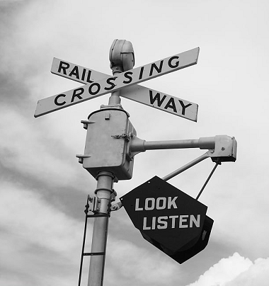 A black-and-white photo of a railroad crossing sign with the words 'RAIL CROSSING' and 'WAY' on the crossing bars. A black sign hanging below reads 'LOOK LISTEN'.