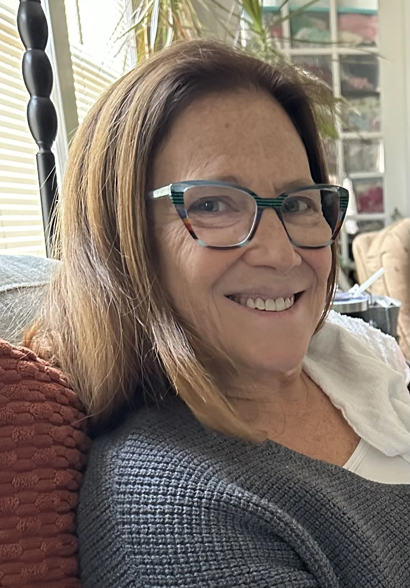 Close-up of a smiling woman with shoulder-length light brown hair, wearing glasses and a gray sweater, sitting on a red textured pillow in a cozy indoor setting with a background of a plant, window blinds, and storage drawers.