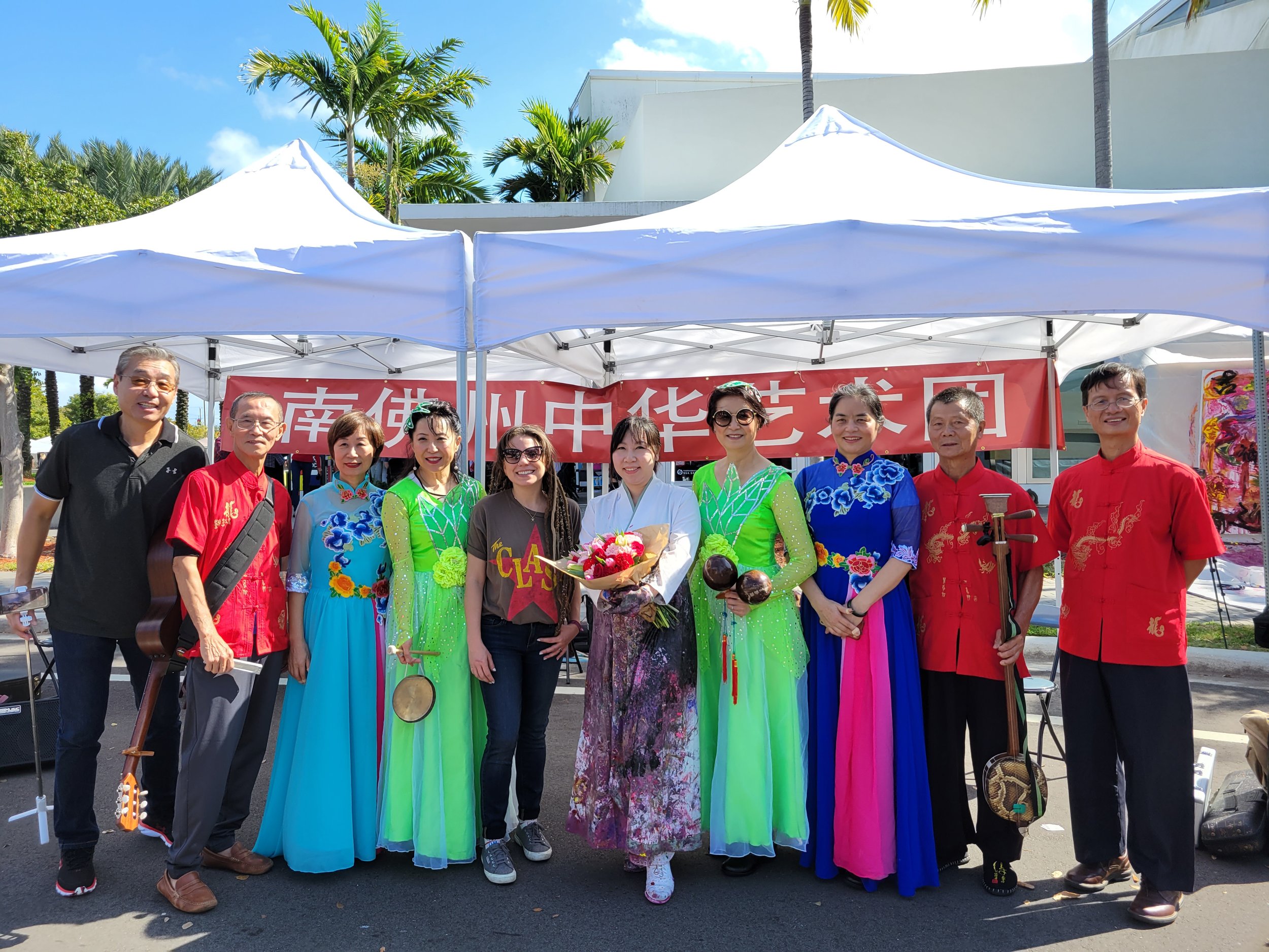 Group of people standing together under a white canopy with a red banner behind them, some in traditional colorful Chinese outfits, outdoors on a sunny day with palm trees.