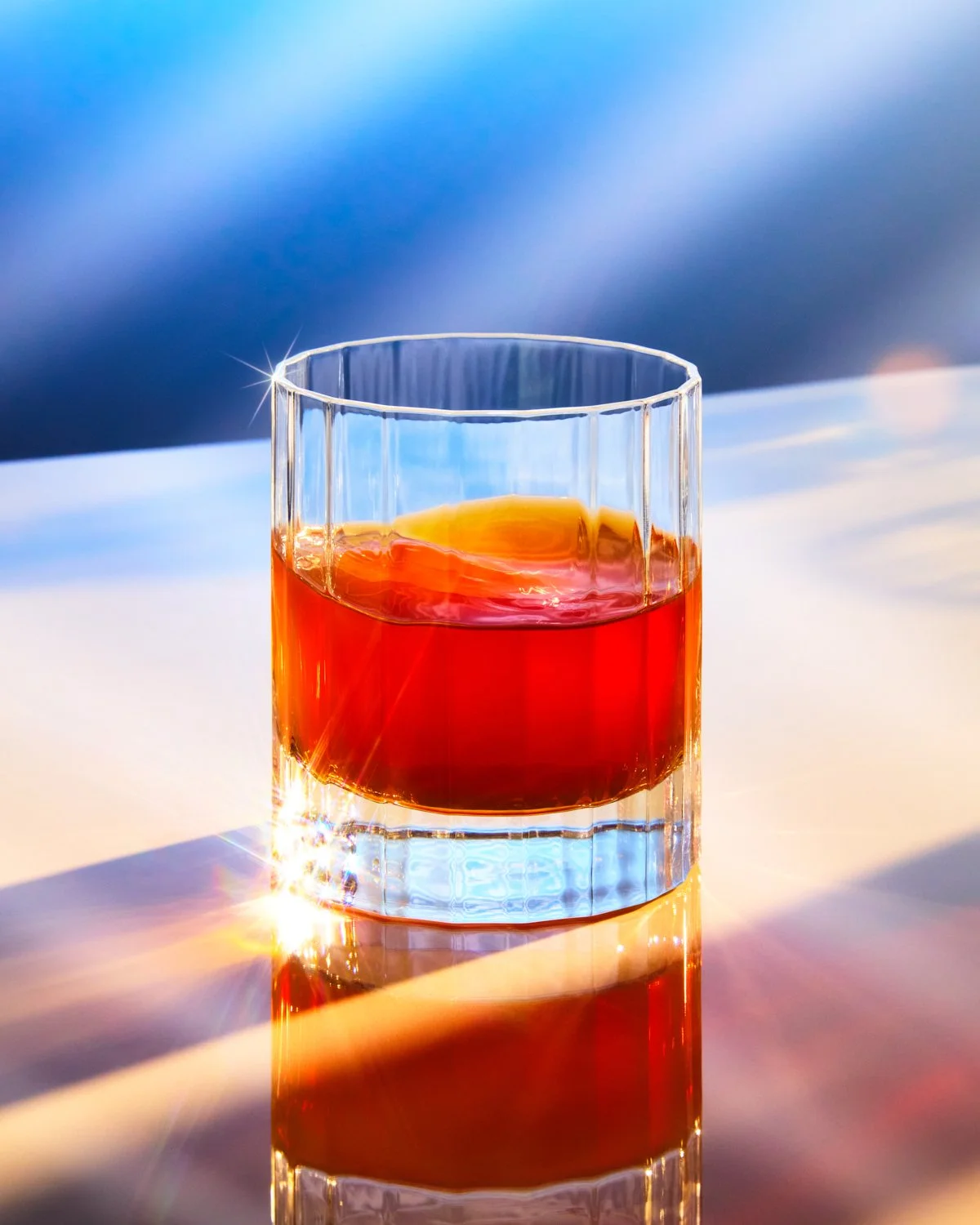 Amber cocktail in a clear rocks glass with dramatic reflections, colorful light, and sparkling highlights on a glossy surface.
