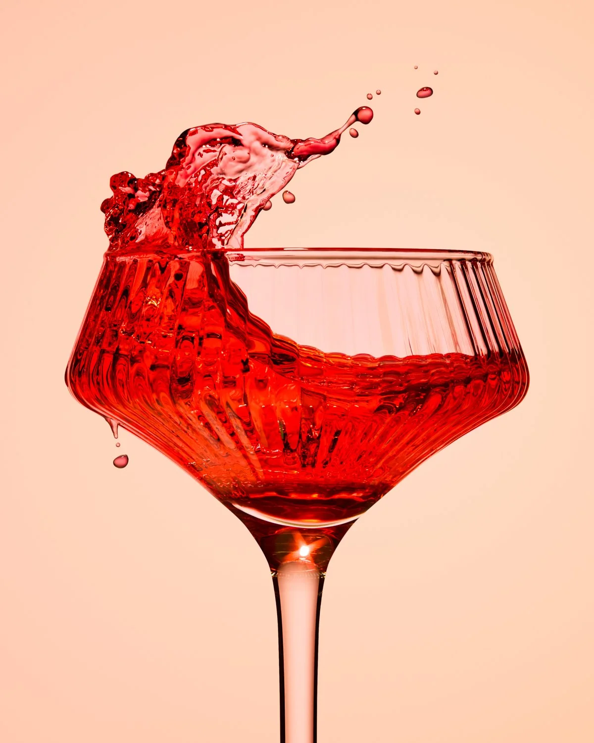 Red cocktail splashing dramatically from a ribbed coupe glass against a warm neutral background, captured with high-speed photography.