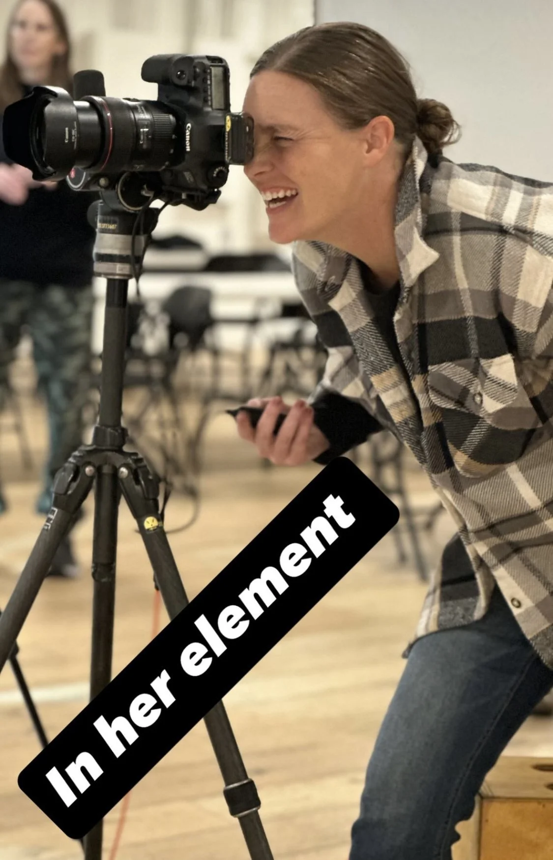 Person looking through a camera viewfinder, smiling, wearing a plaid shirt, in a room with chairs and standing woman in the background, with a black text overlay that says 'In her element.'