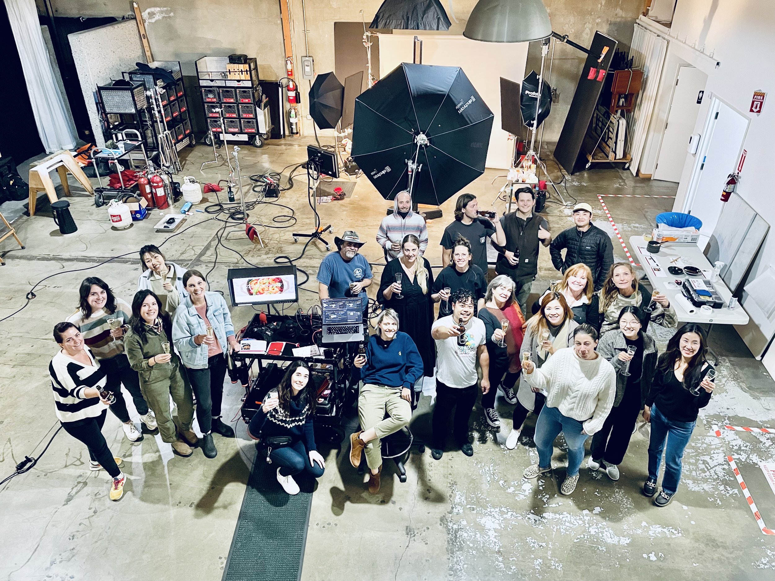 Group of people gathered in a photography or video studio, celebrating with drinks, some holding glasses or phones, with professional lighting, camera equipment, and backdrop in the background.