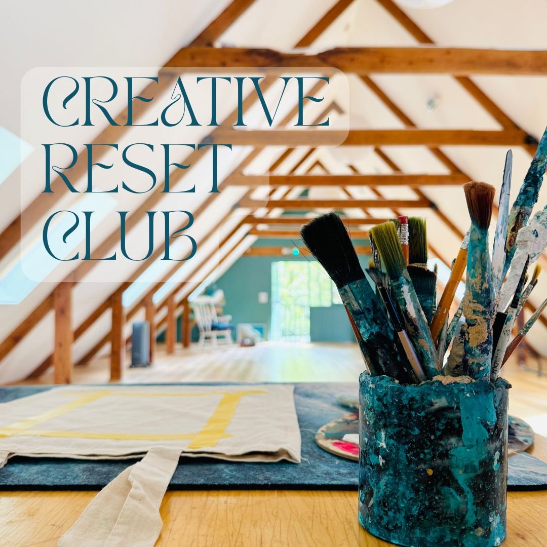 Interior view of an attic studio with wooden beams, featuring a collection of paintbrushes in a paint-splattered container in the foreground, and a quilt on the table. Text overlay reads 'Creative Reset Club'.