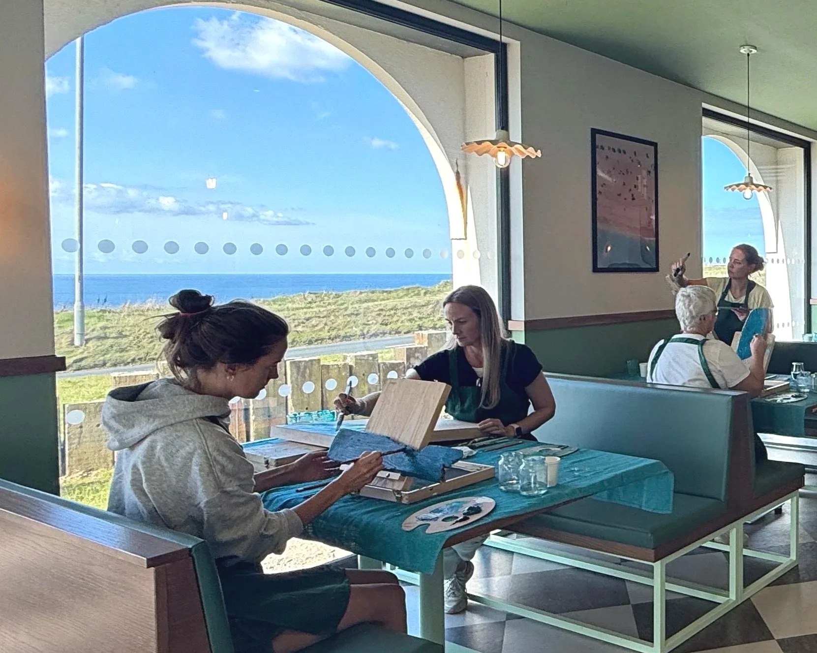 People sitting inside a restaurant with large windows overlooking a scenic ocean view. Two women are painting on small canvases, and a woman in the background is taking a photo. The interior features hanging lights and framed artwork on the walls.