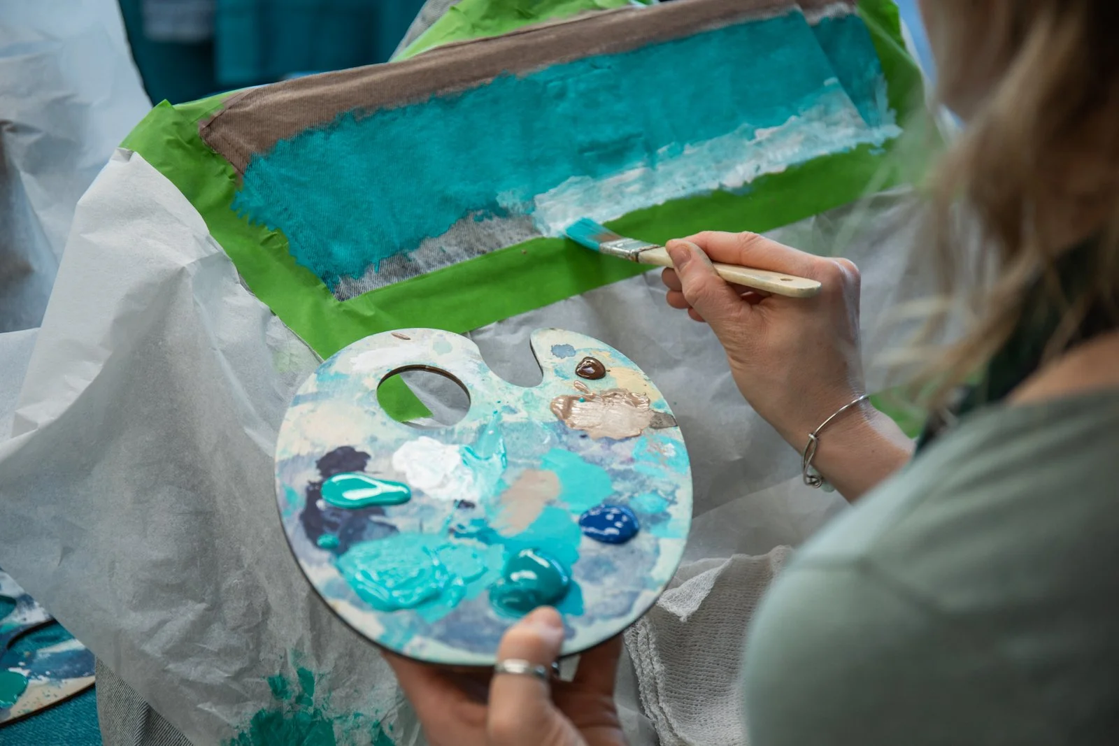 Person painting on a canvas with various shades of blue, green, and beige using a paintbrush, holding a palette with multiple colors of paint.