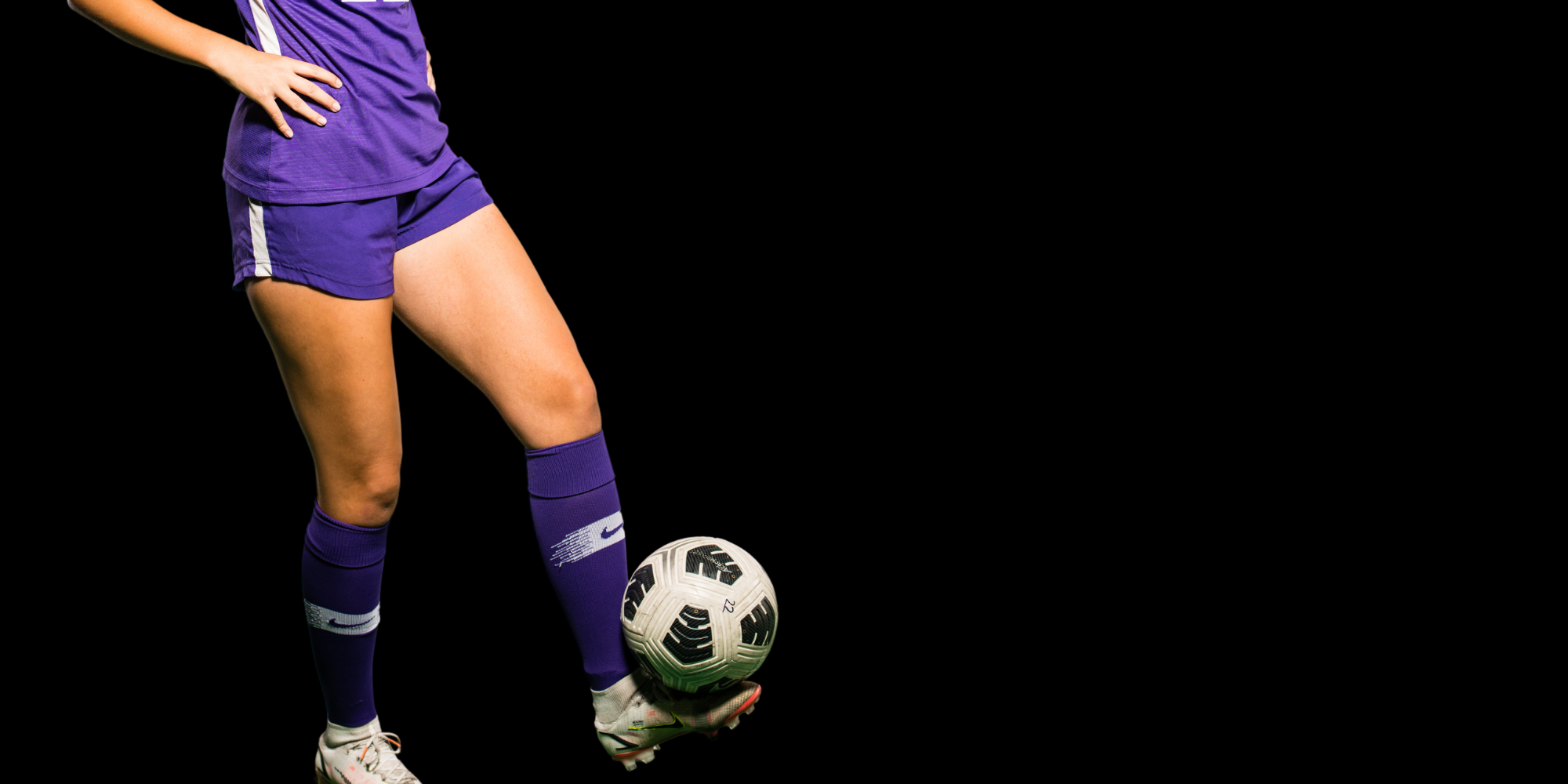 Foot of female soccer player in purple uniform balancing soccer ball on her foot against black background