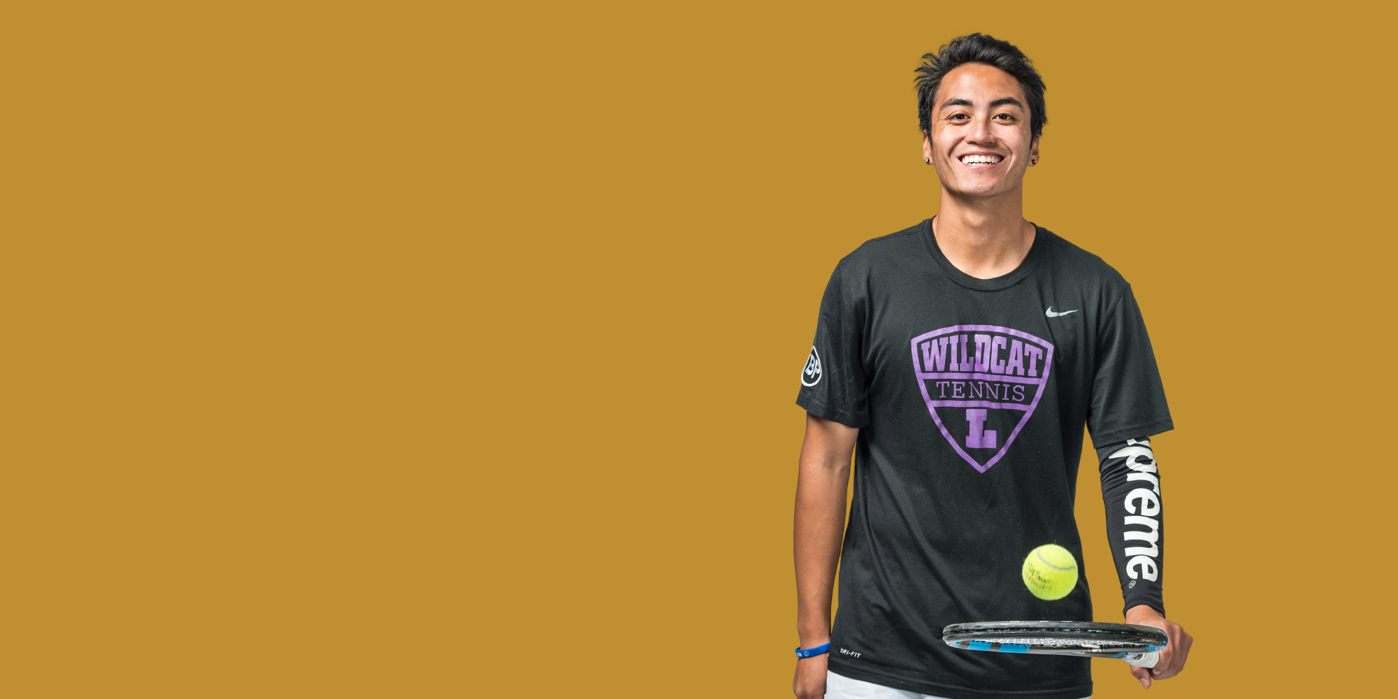 A young man with dark hair smiling, wearing a black tennis practice shirt with purple 'Wildcat Tennis' logo, holding a tennis racket with a tennis ball balanced on it, standing against a plain mustard-colored background.