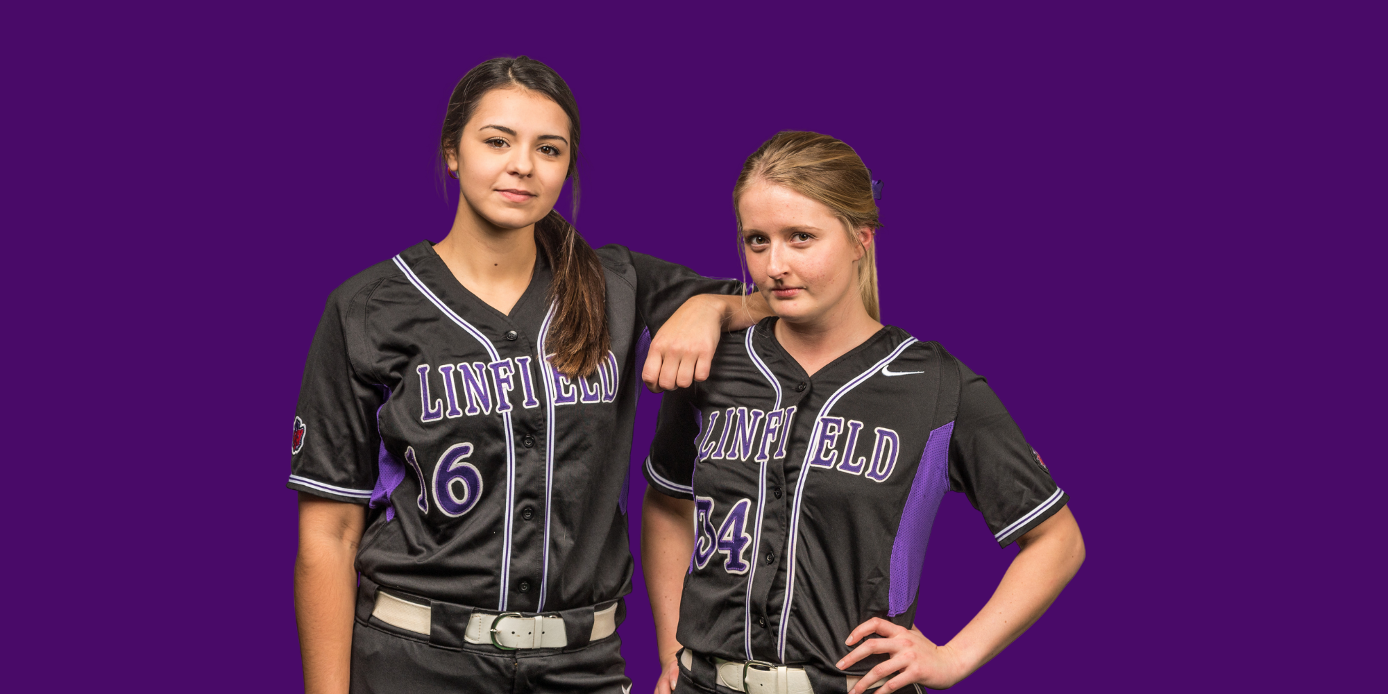 Two young female softball players in black jerseys with purple and white accents, standing against a purple background. One has her arm over the other's shoulder.