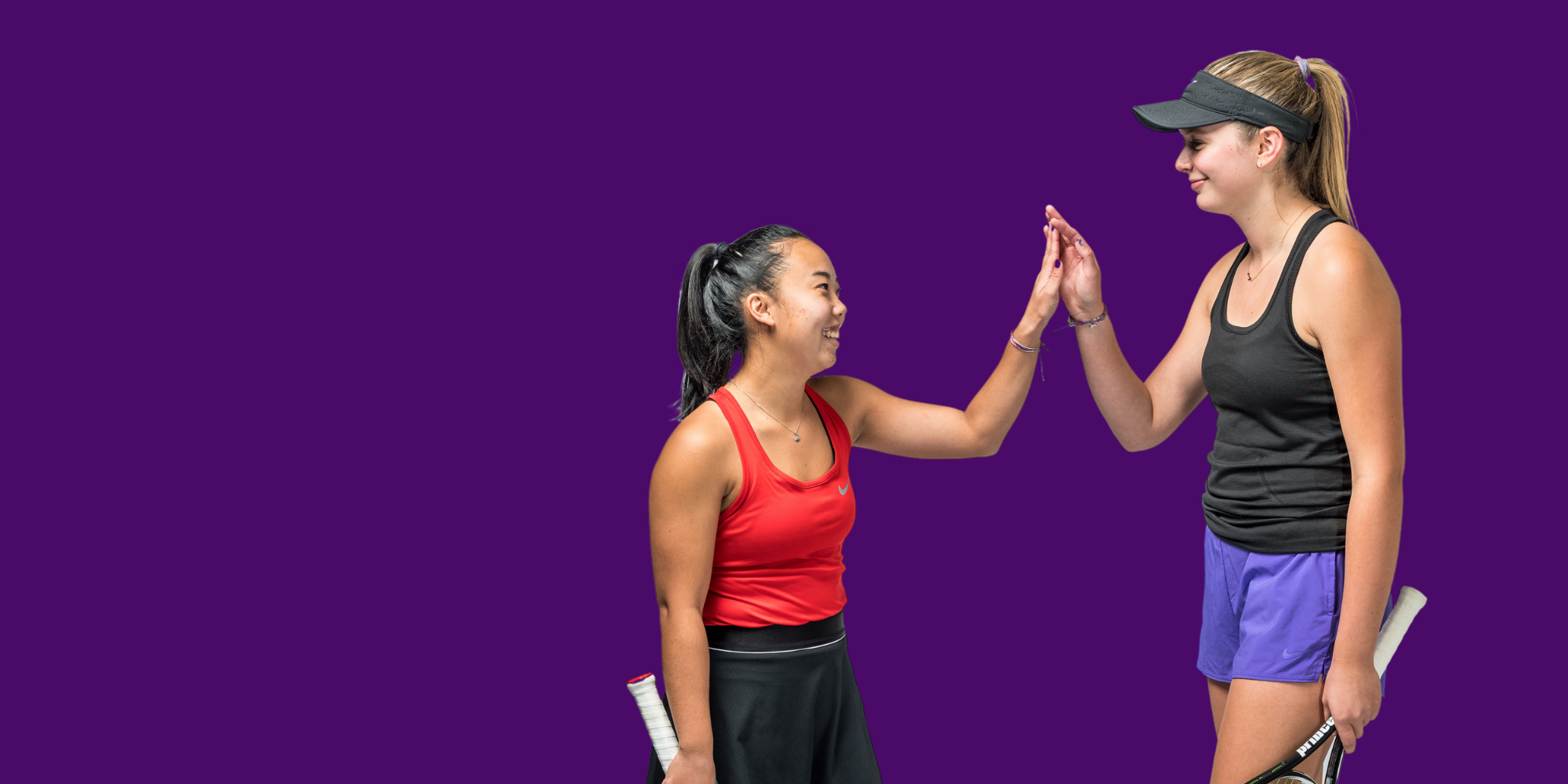 Two young female tennis players, one in a red tank top and black shorts, the other in a black tank top and purple shorts, high-fiving each other after a game against a purple background.