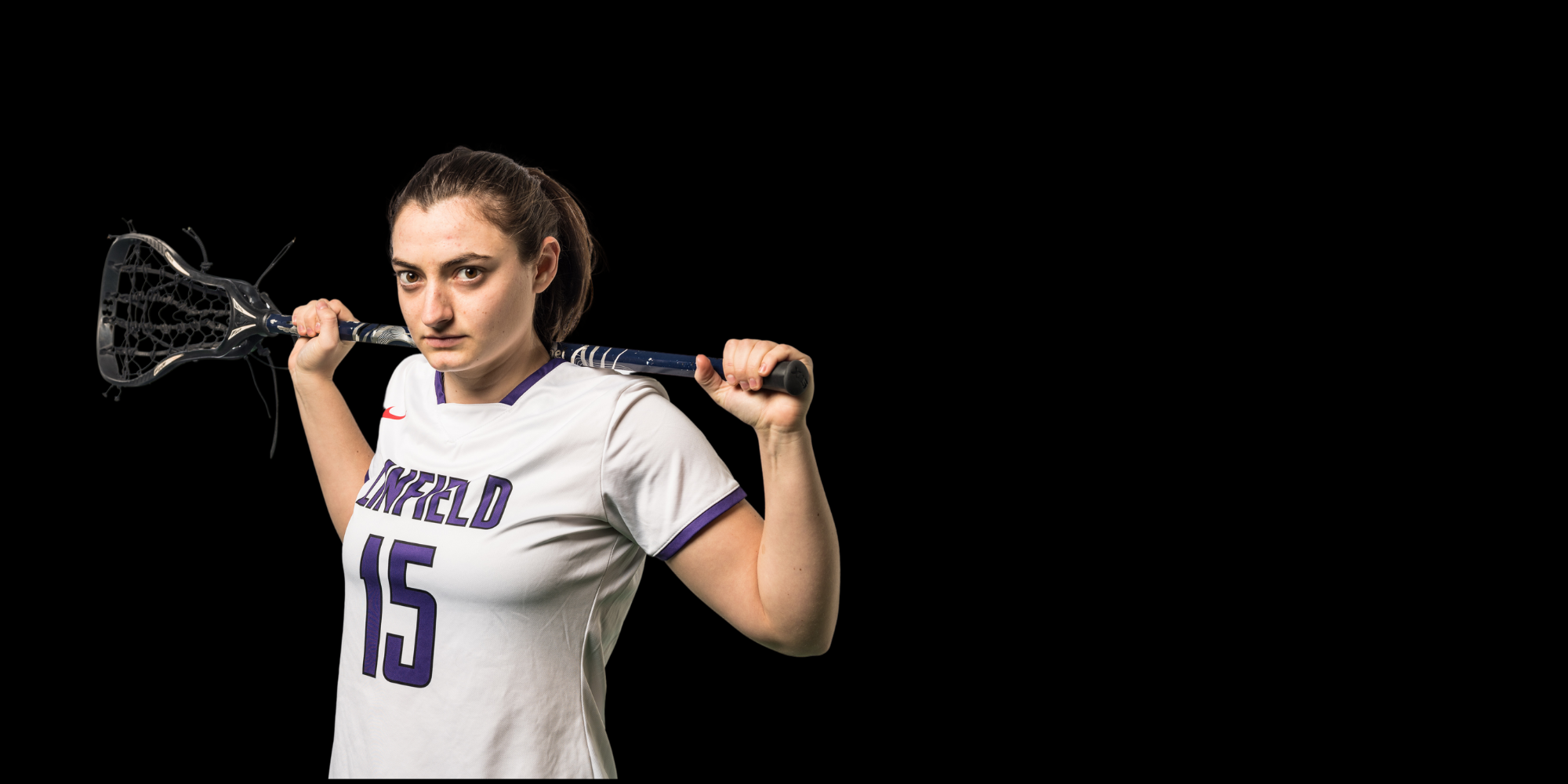 A female Linfield University lacrosse player holding a lacrosse stick over her shoulders and standing against a black background.