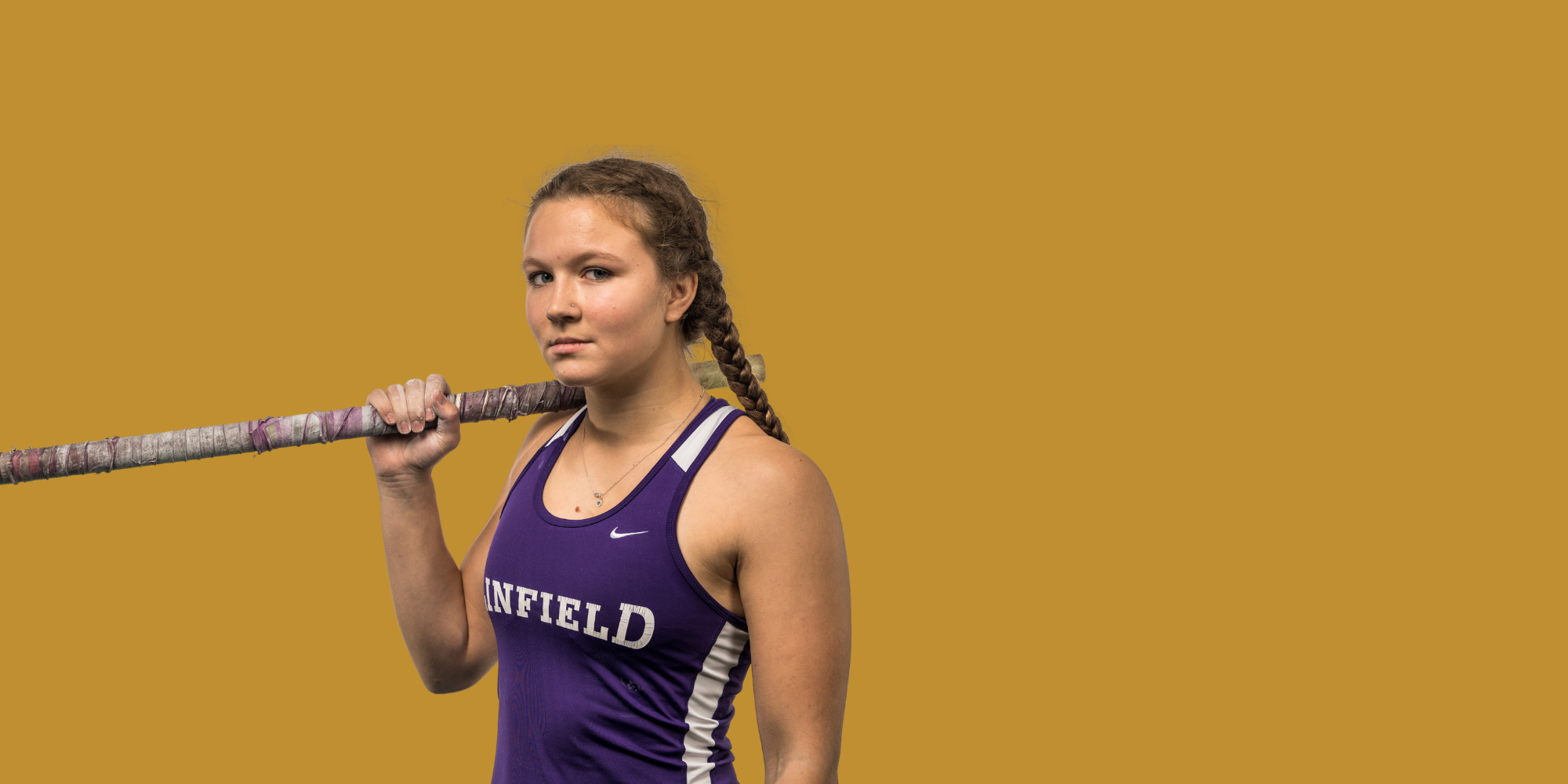 A young female athlete with braided hair, wearing a purple track sports uniform with the word 'LINFIELD' on it, holding a pole over her shoulder, standing against a solid mustard yellow background.