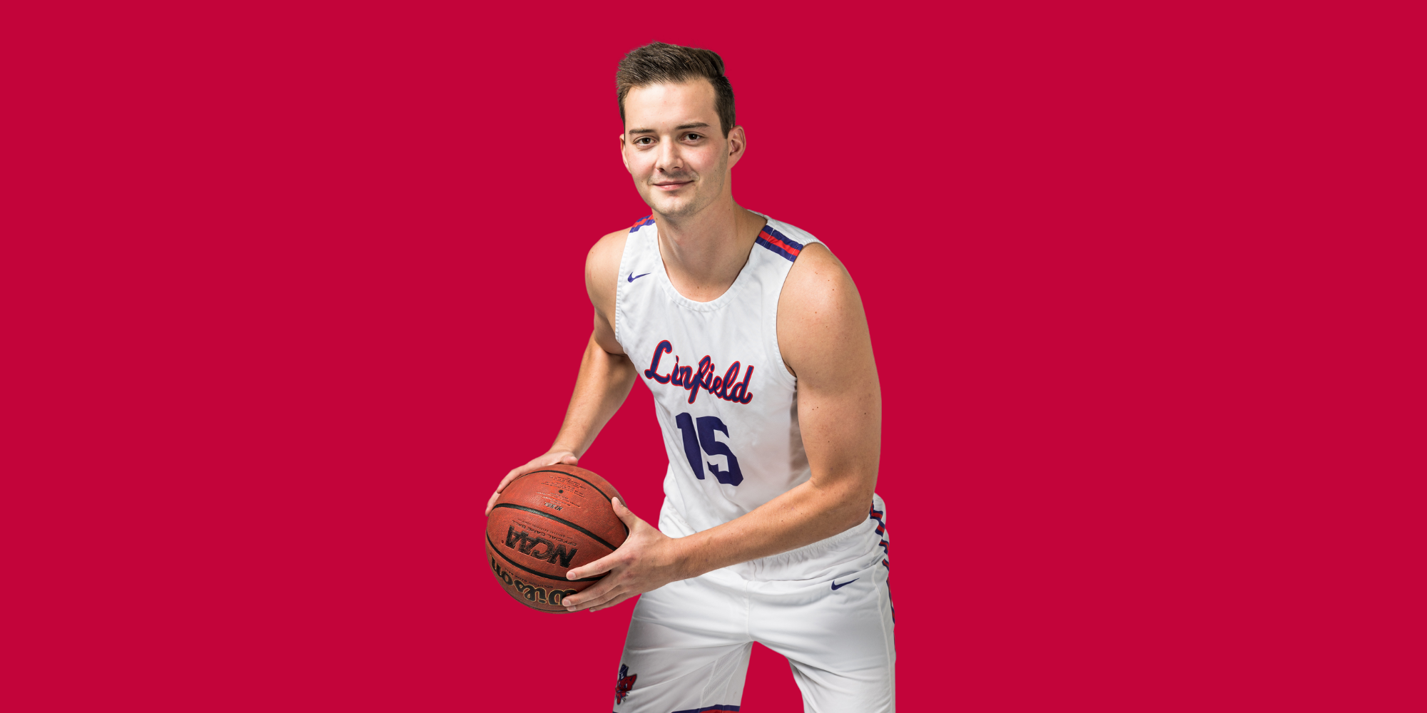 Young male basketball player in white uniform with blue and red accents holding a basketball, standing against a solid red background.