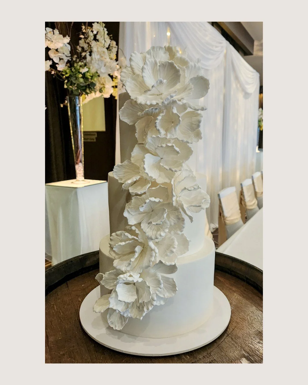 A breathtaking three-tier white fondant cake adorned with a full deconstructed floral drape for Flora &amp; Evan, layered with chocolate and white chocolate berry flavours for a timeless, romantic finish. ✨

TASTEMONIAL
&ldquo;We&rsquo;d just like to
