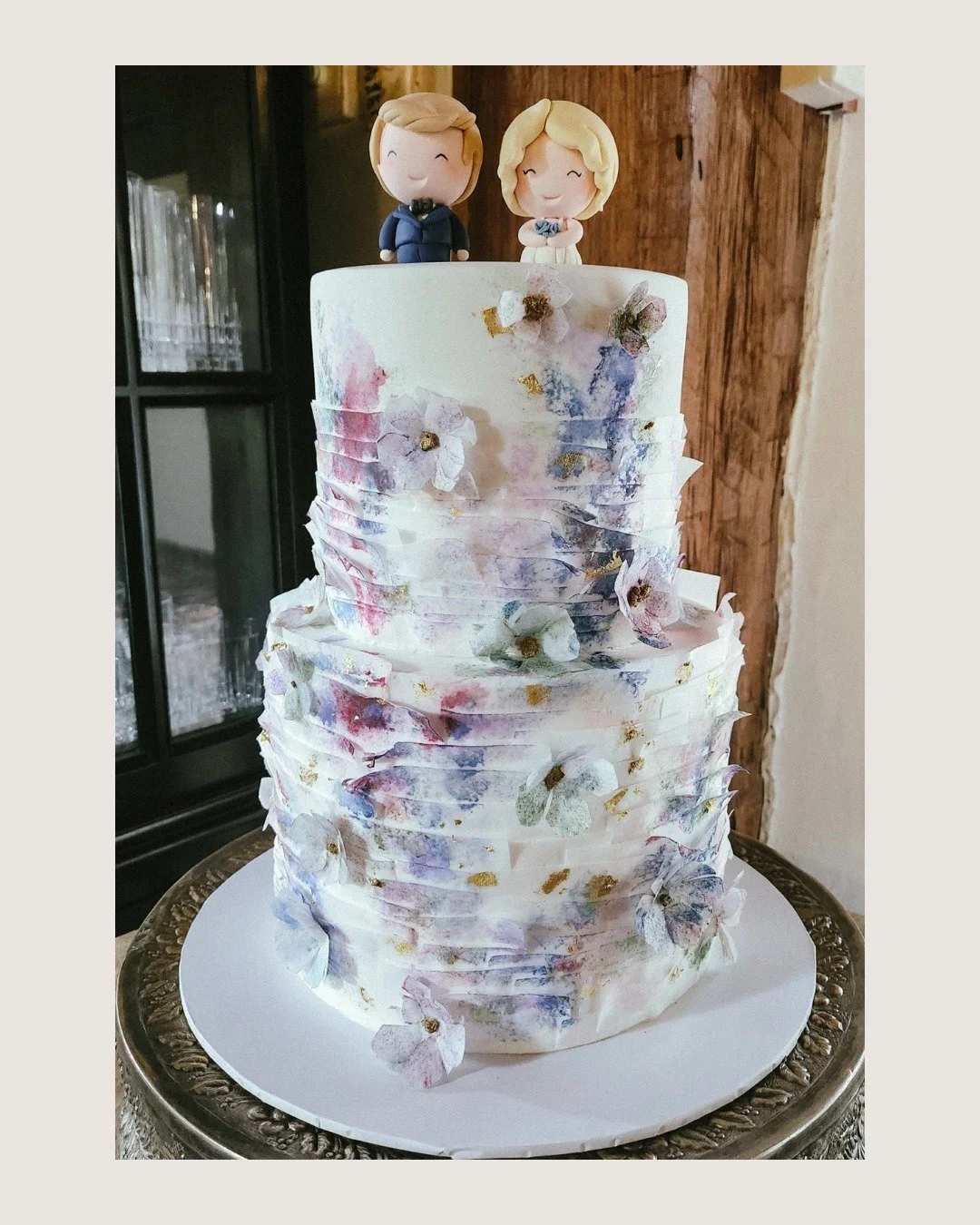A dreamy two-tier design featuring white chocolate and chocolate, wrapped in white fondant with square overlap ruffles, full coverage on the base tier and soft half coverage above.

Finished with delicate blue watercolour detailing, flecks of gold le