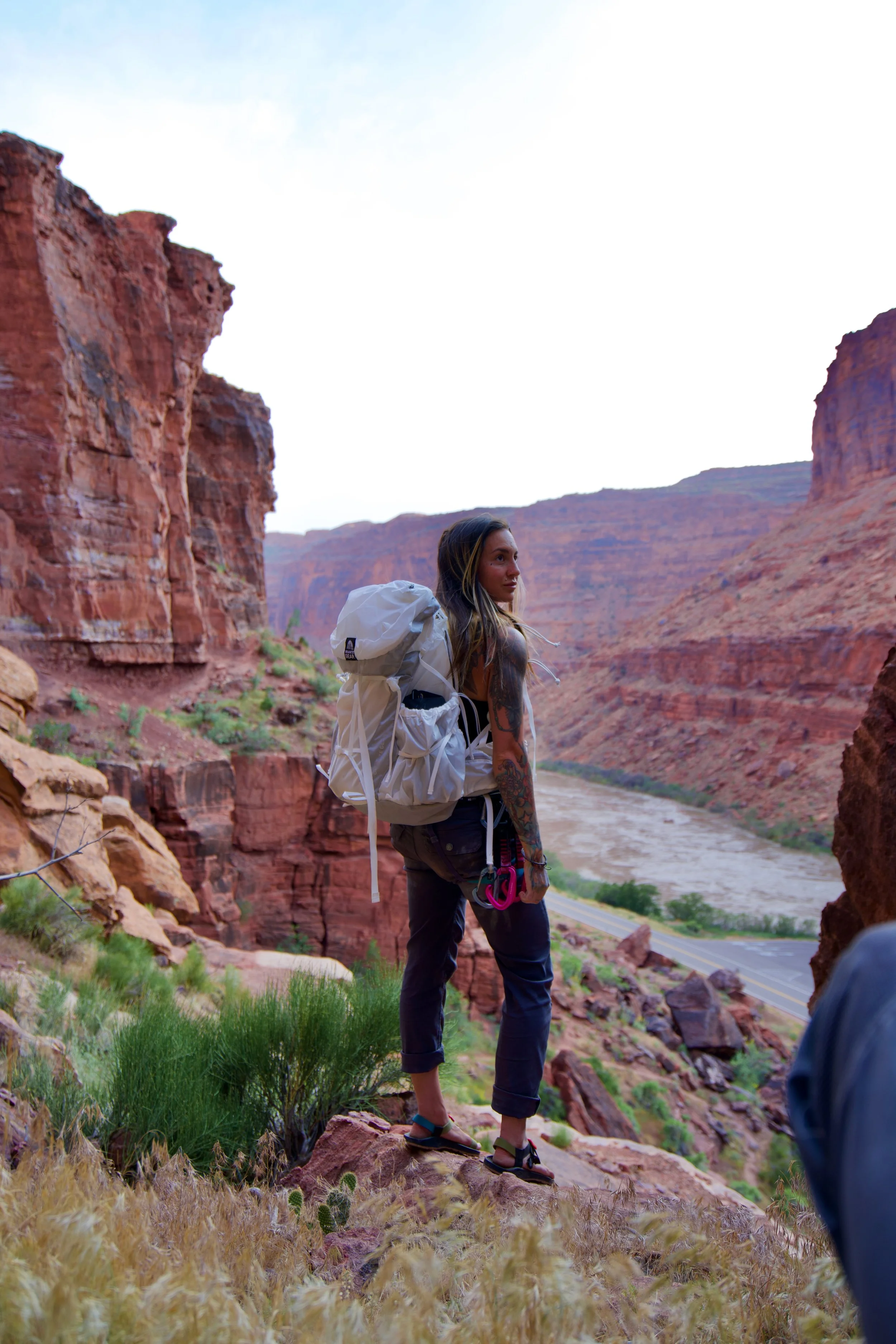 Female wellness and outdoor industry copywriter wearing white backpack stands on sandstone cliff above a river near Moab, Utah.