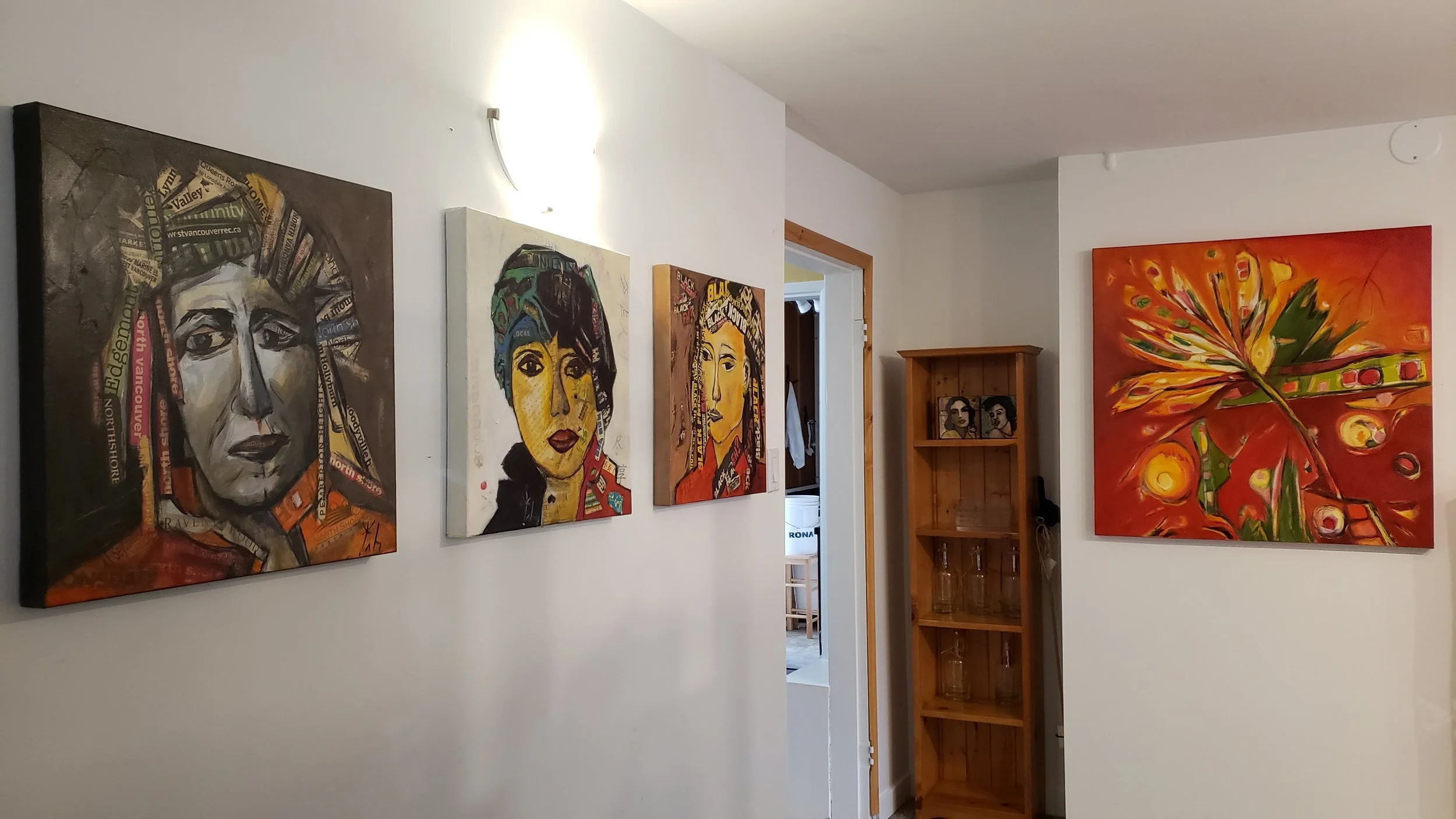 Gallery wall with four colorful portraits and a large abstract painting on white walls, wooden shelving unit in background.