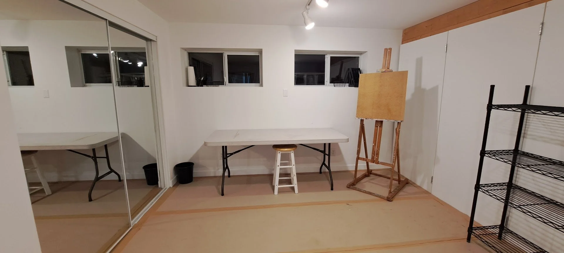 Empty art studio with three small windows, a white folding table, a wooden easel with a canvas, a stool, a black metal shelving unit, and a large mirror closet door.