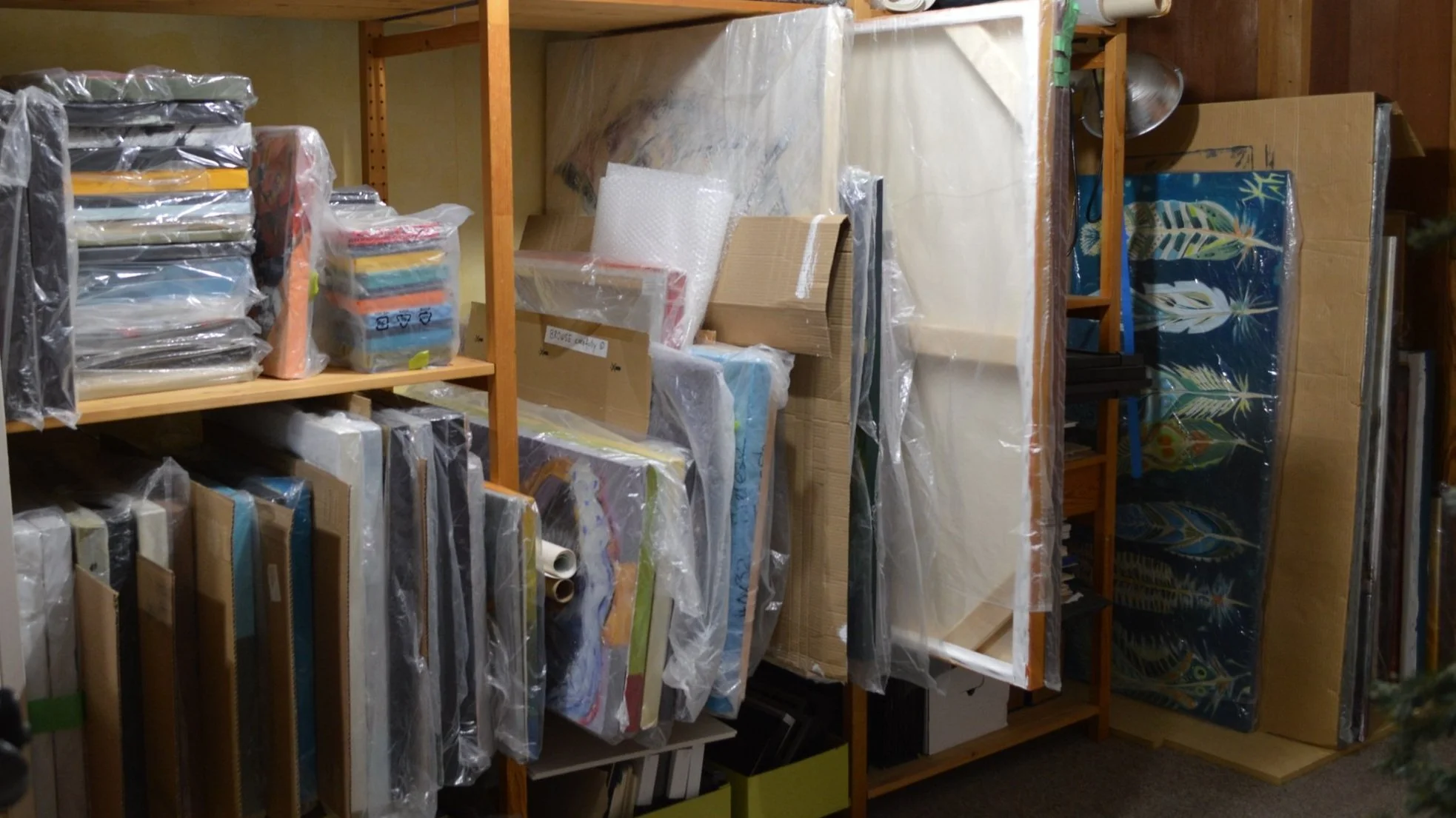 Storage room with shelves filled with wrapped canvases, art supplies, and packages
